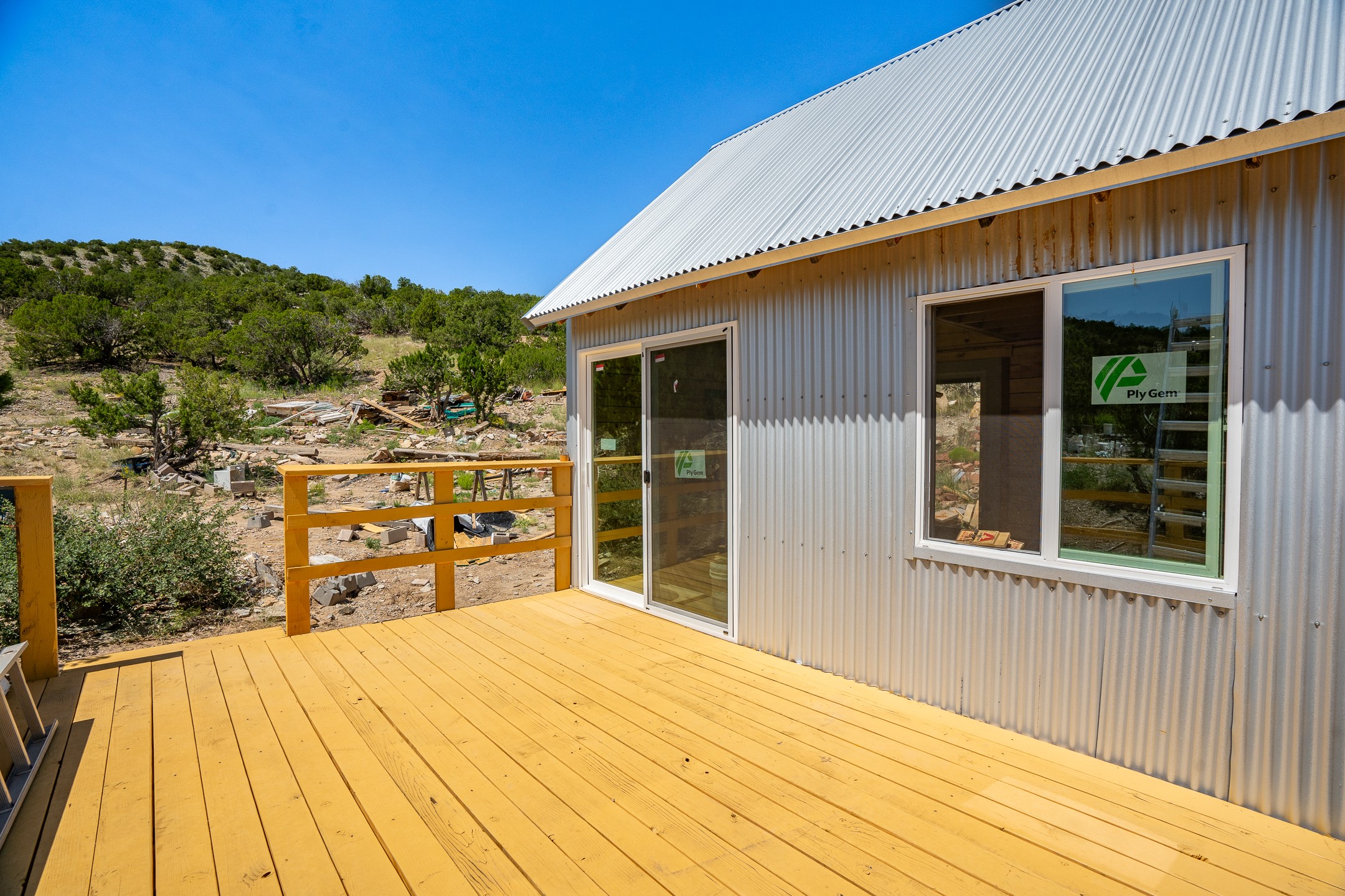 New wooden deck beside a corrugated metal home, with sliding doors and a scrub-covered hillside beyond.