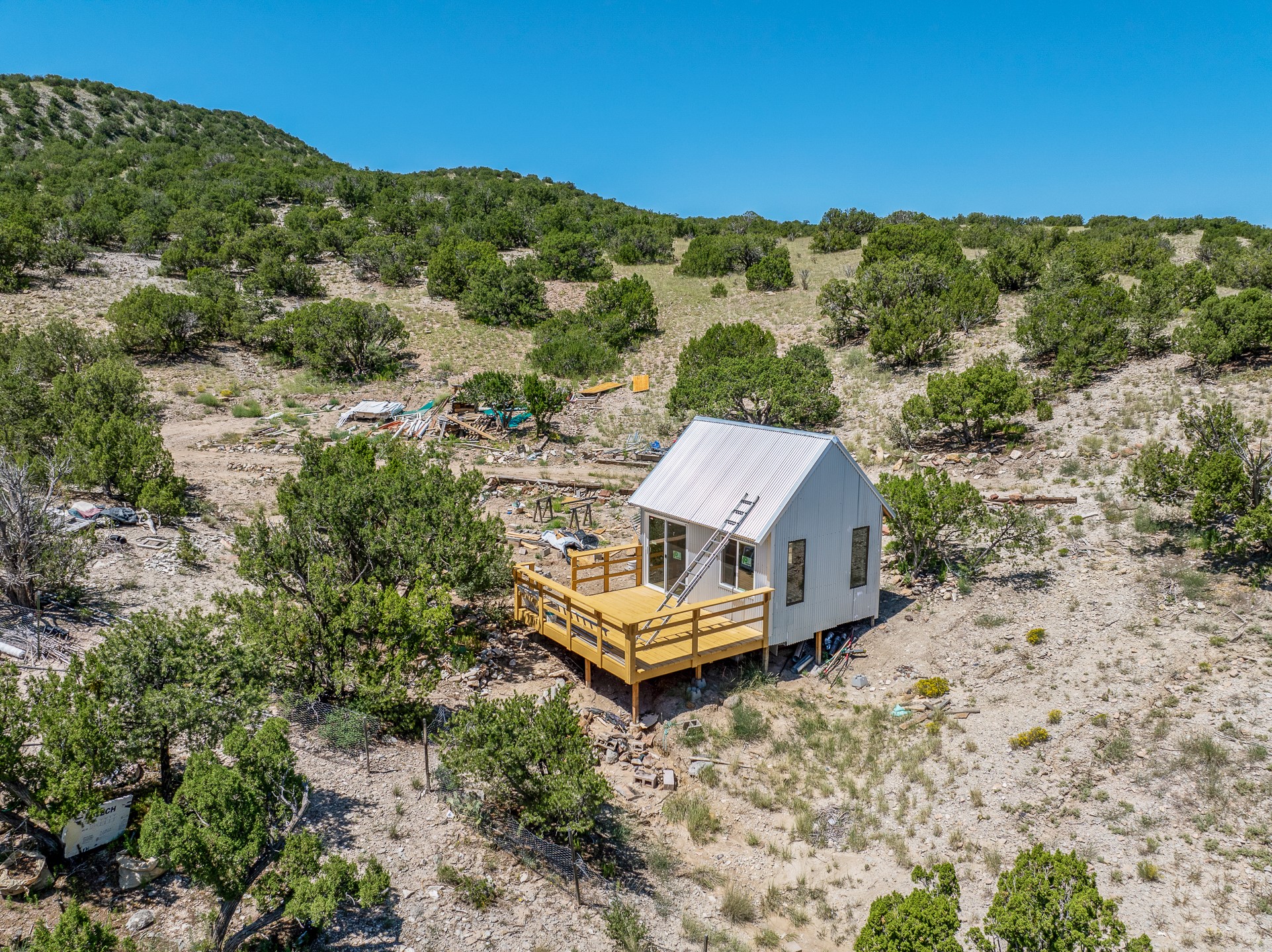 Small white cabin with a wooden deck on a rocky, shrub-covered hillside, seen from above.