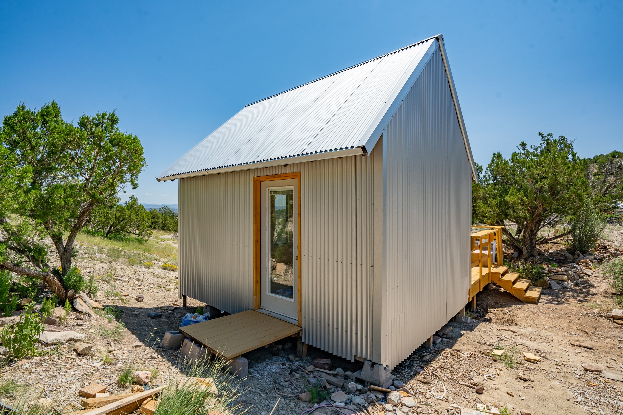 Small metal-sided cabin with a steep white roof and wooden deck on rocky terrain among trees.
