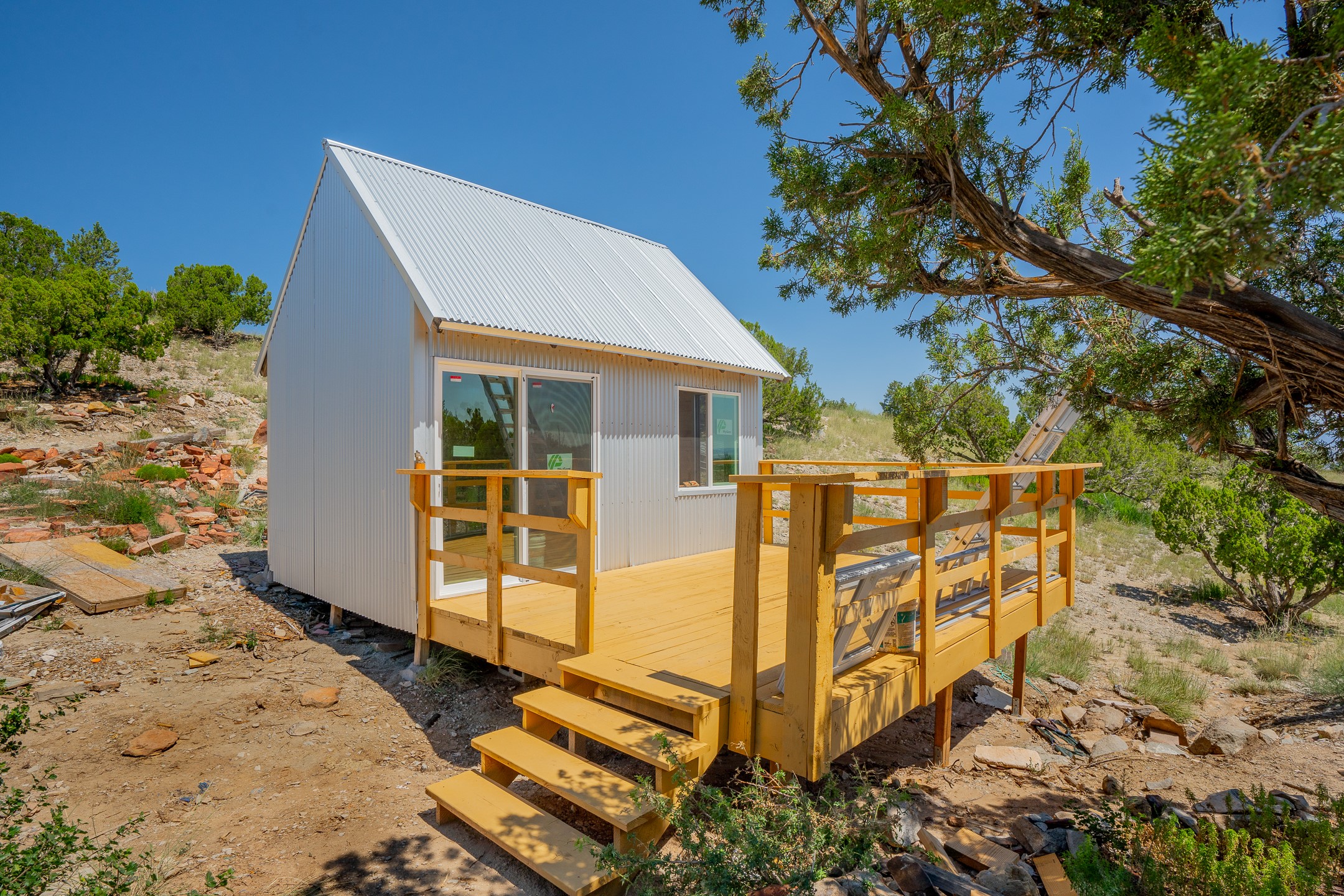 Small white metal cabin with a steep roof and new wooden deck on a rocky hillside.