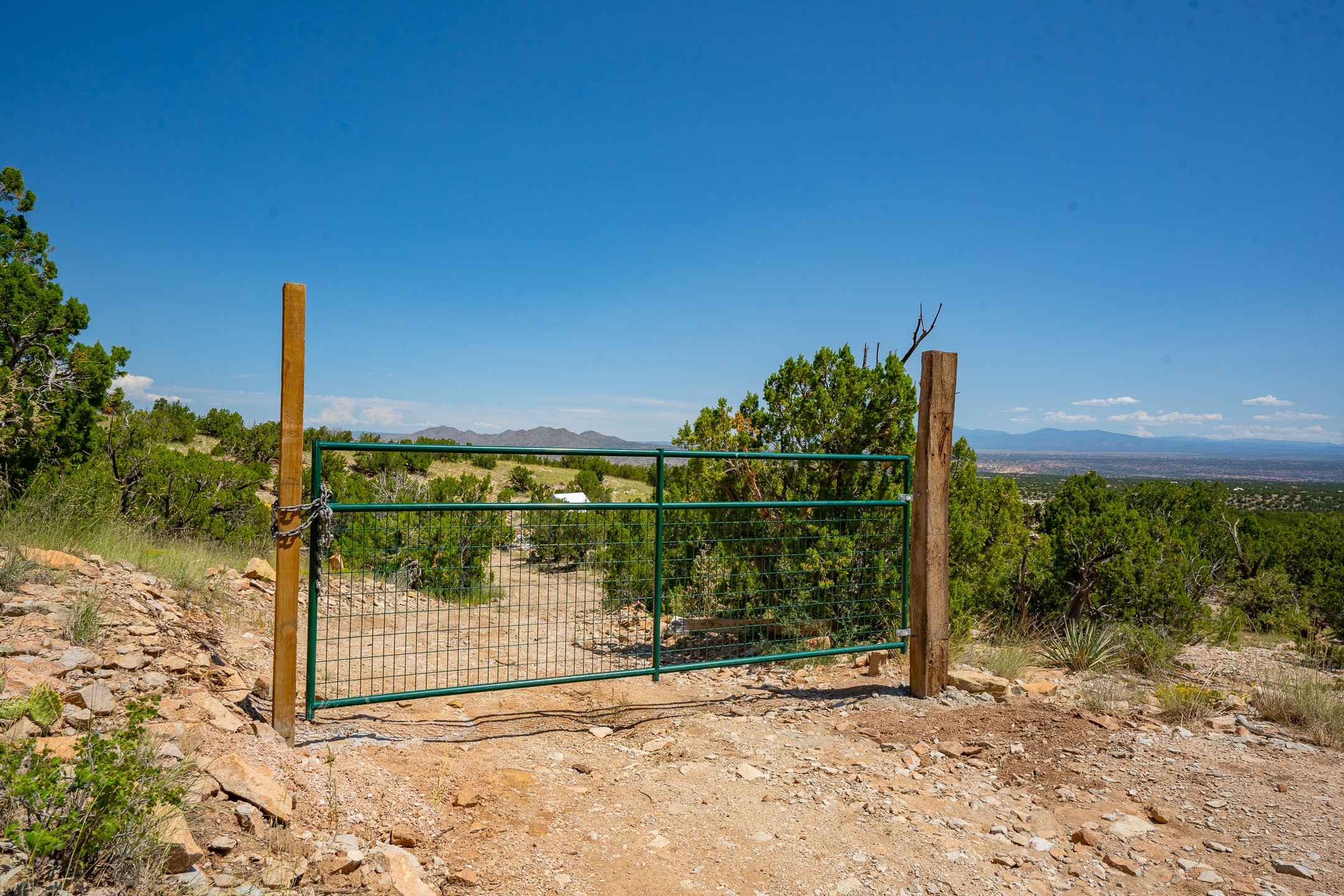 Green metal gate at a rocky rural property entrance, with scrubland and distant mountain views.