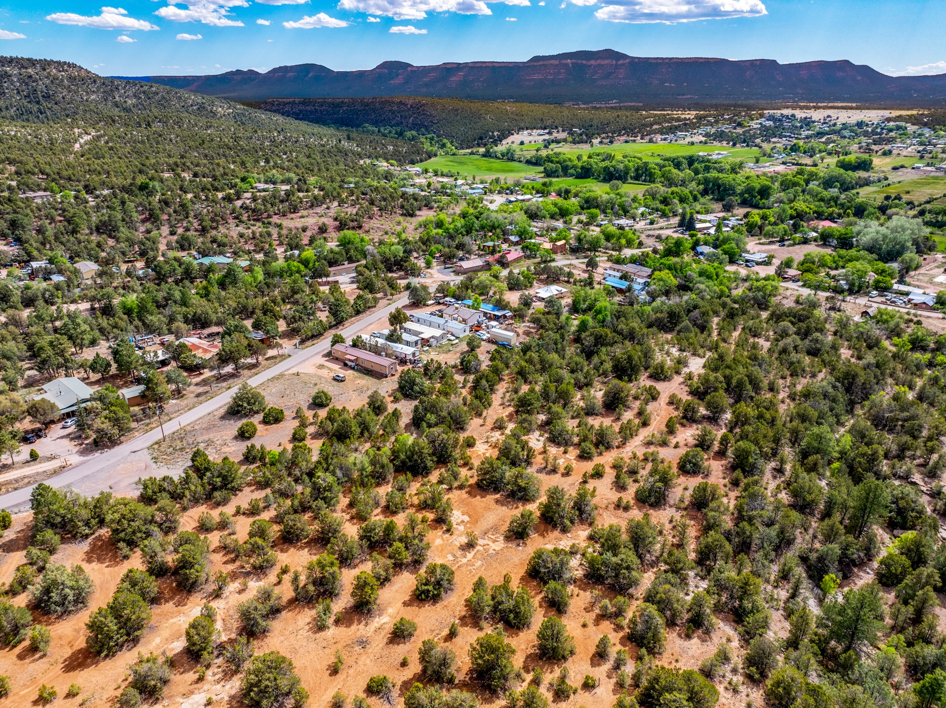 Aerial view of shrub-covered land beside a rural road, with homes, green fields, and distant mesas.