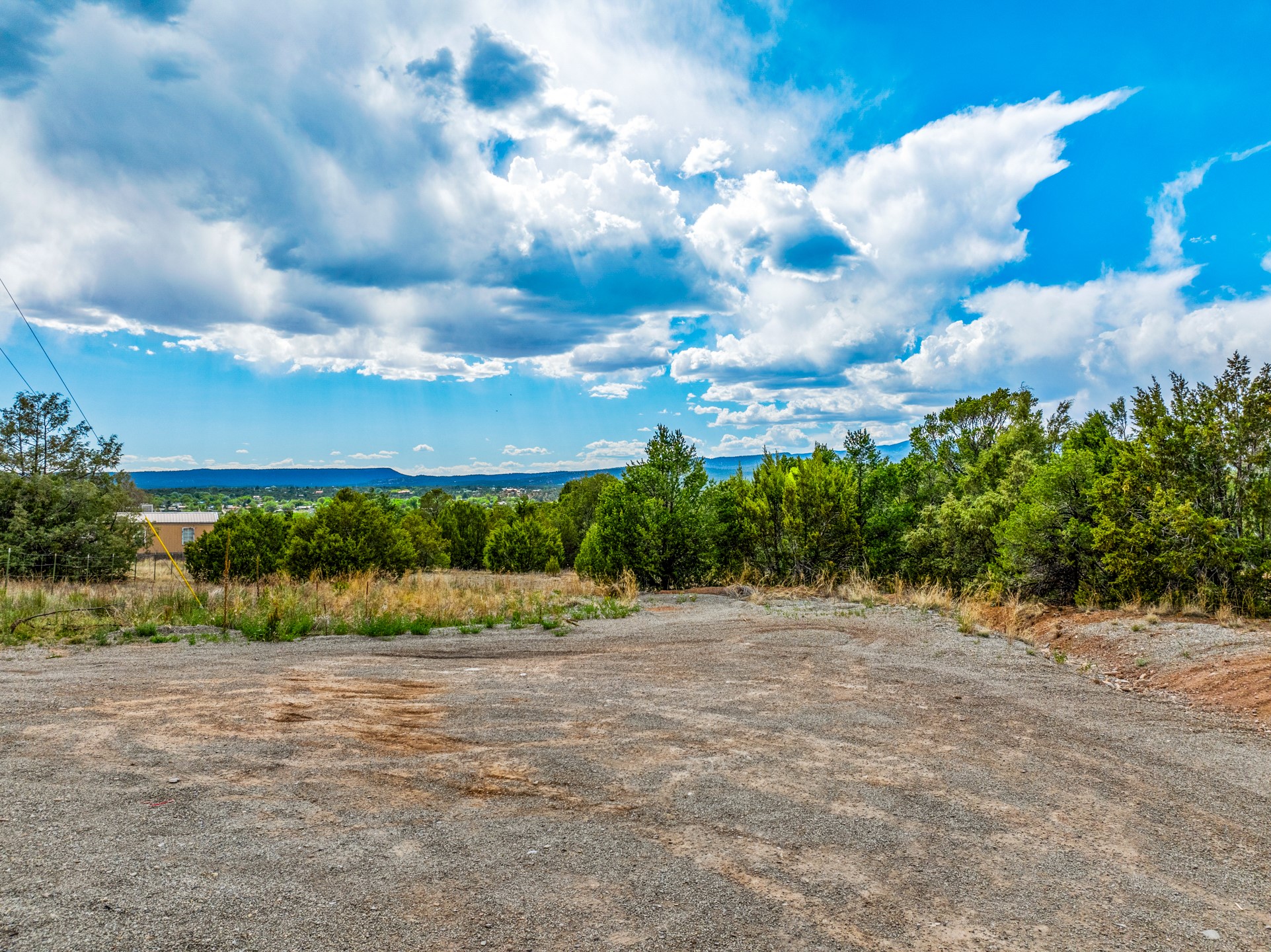 Vacant land with a gravel clearing, tree-lined edges, and distant hills beneath a cloudy sky.