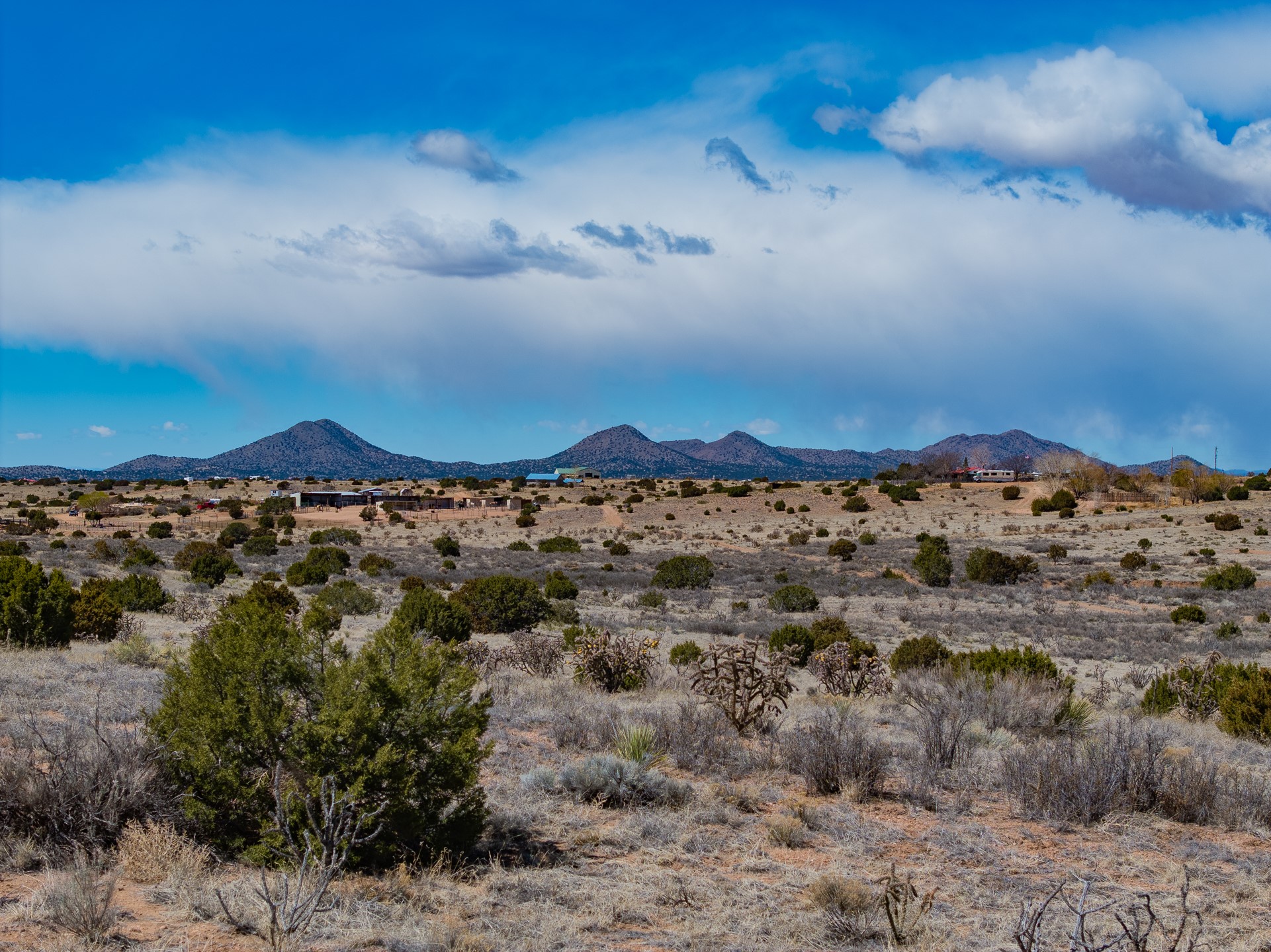 Dry scrubland with scattered shrubs and distant mountain views beneath a dramatic cloud-filled sky.