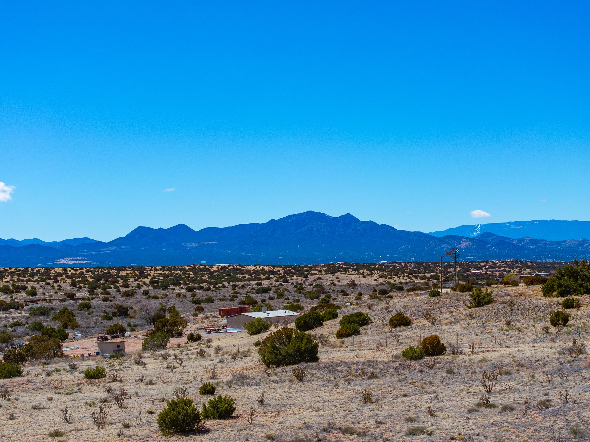 Dry acreage with scattered shrubs, small buildings, and a windmill, with mountains in the distance.