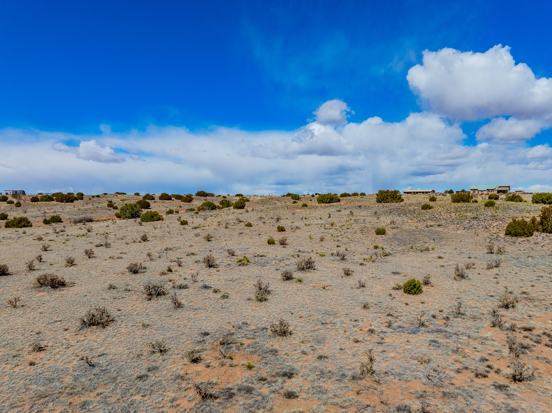 Dry, gently rolling acreage with sparse shrubs and distant buildings beneath a cloud-filled blue sky.