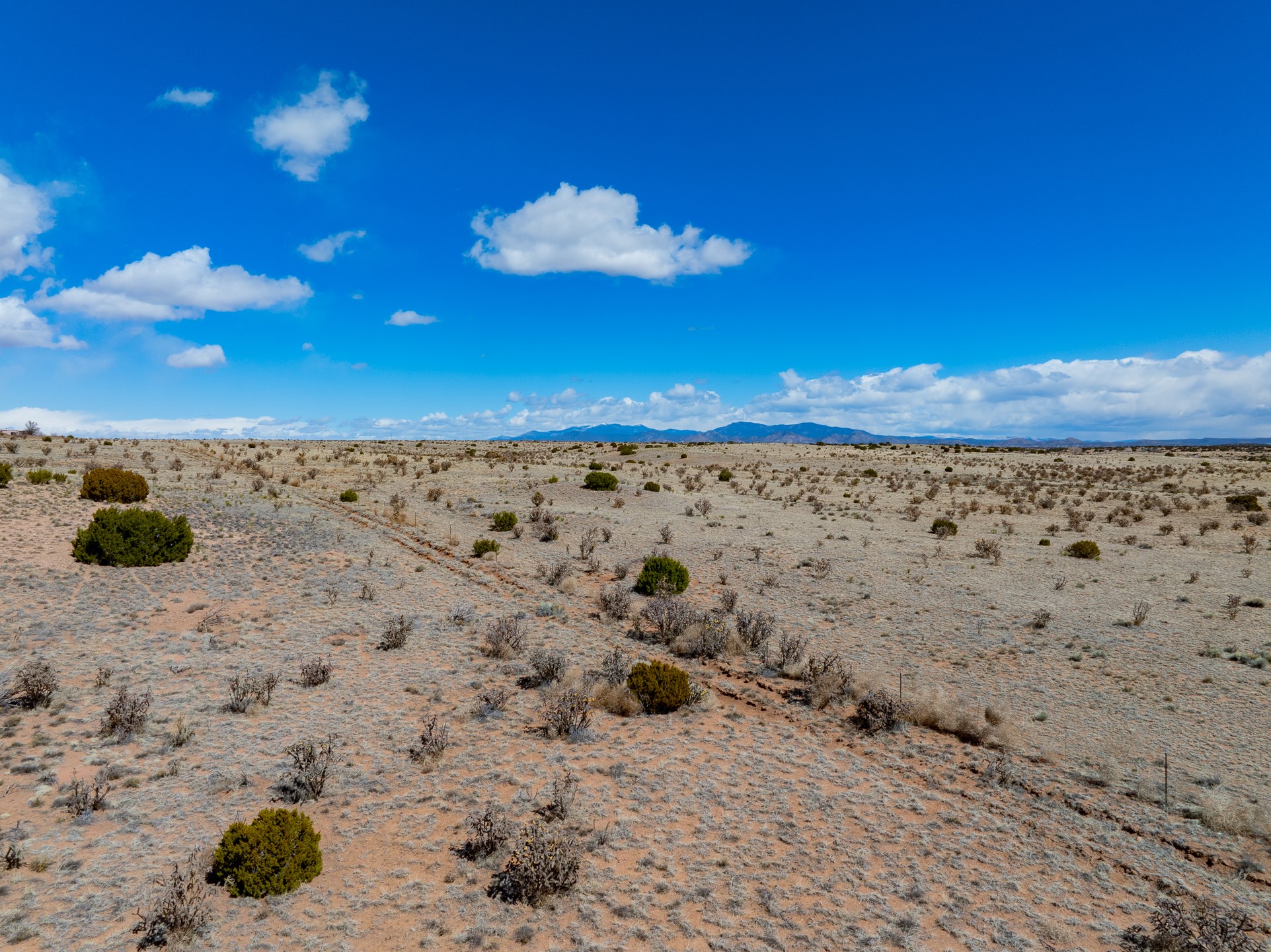 Open desert acreage with scattered shrubs and distant mountains beneath a cloud-filled blue sky.