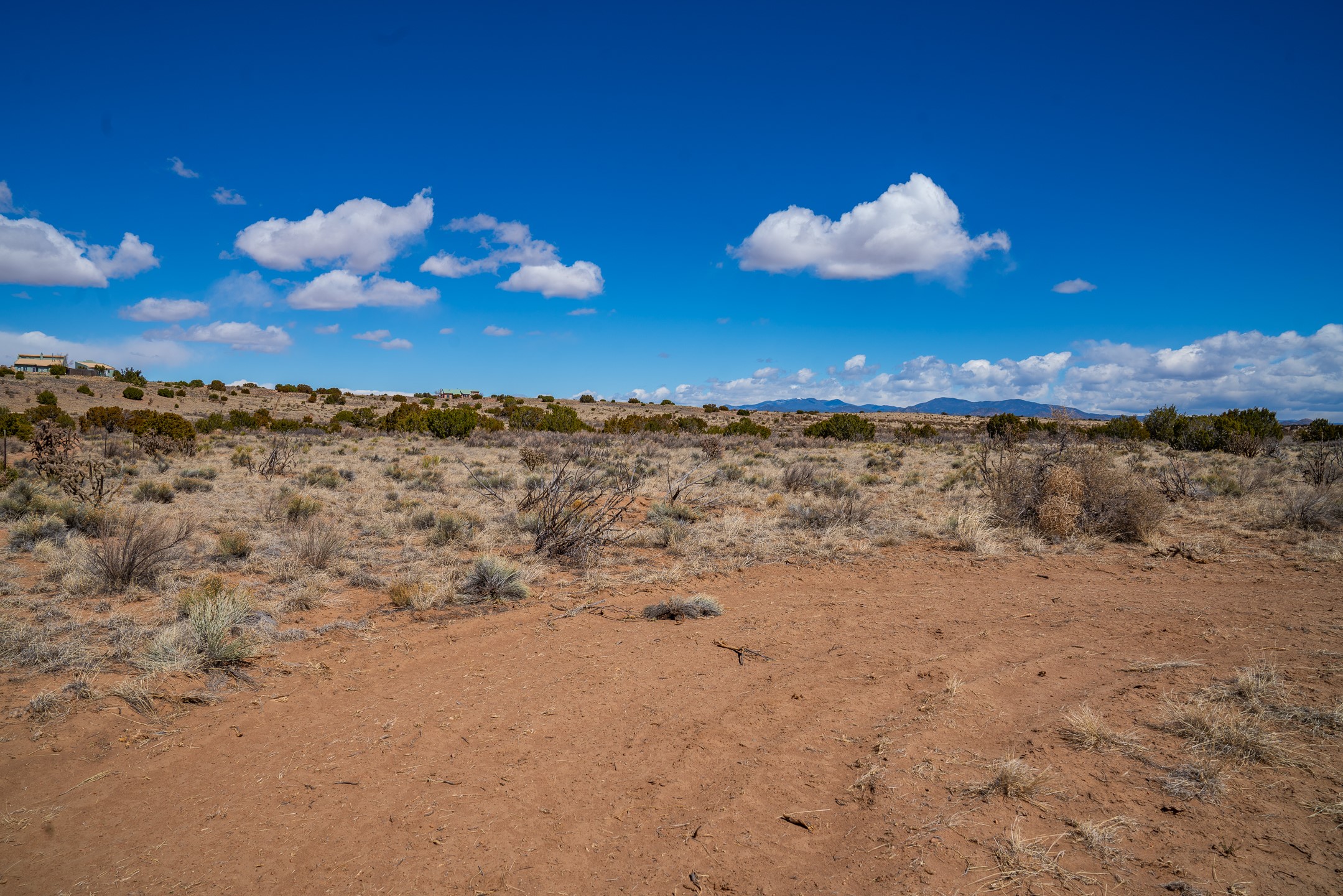 Open scrubland with reddish soil, distant homes, and mountains beneath a blue sky.