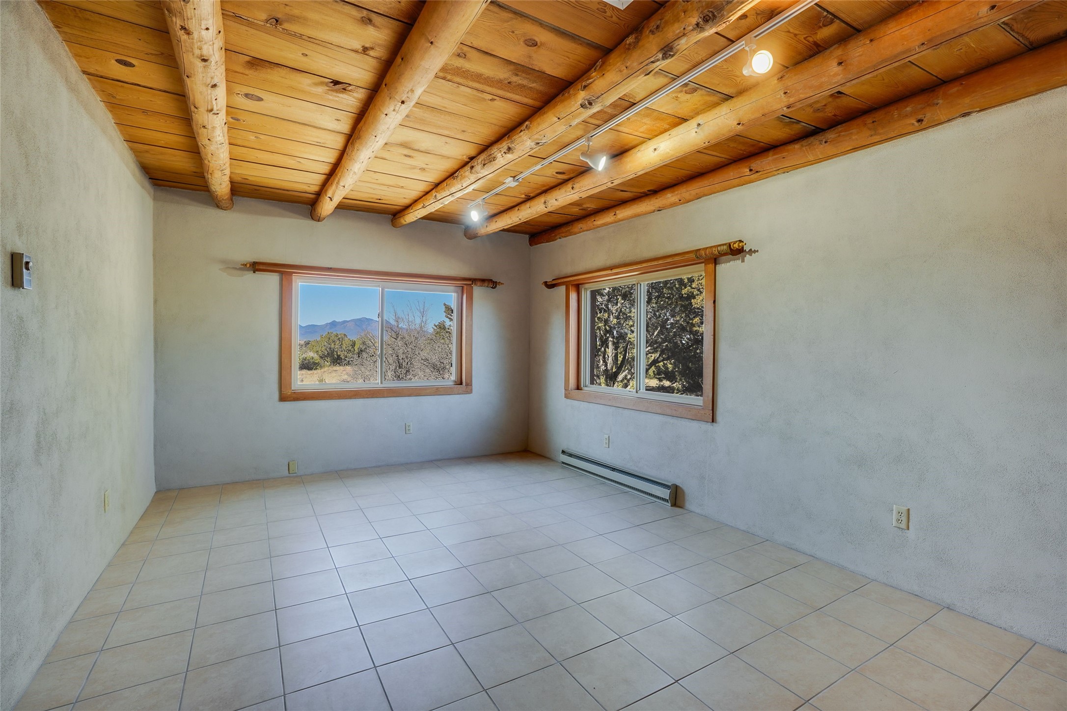 Empty room with exposed timber ceiling beams, tiled floor, and windows overlooking trees and mountains.