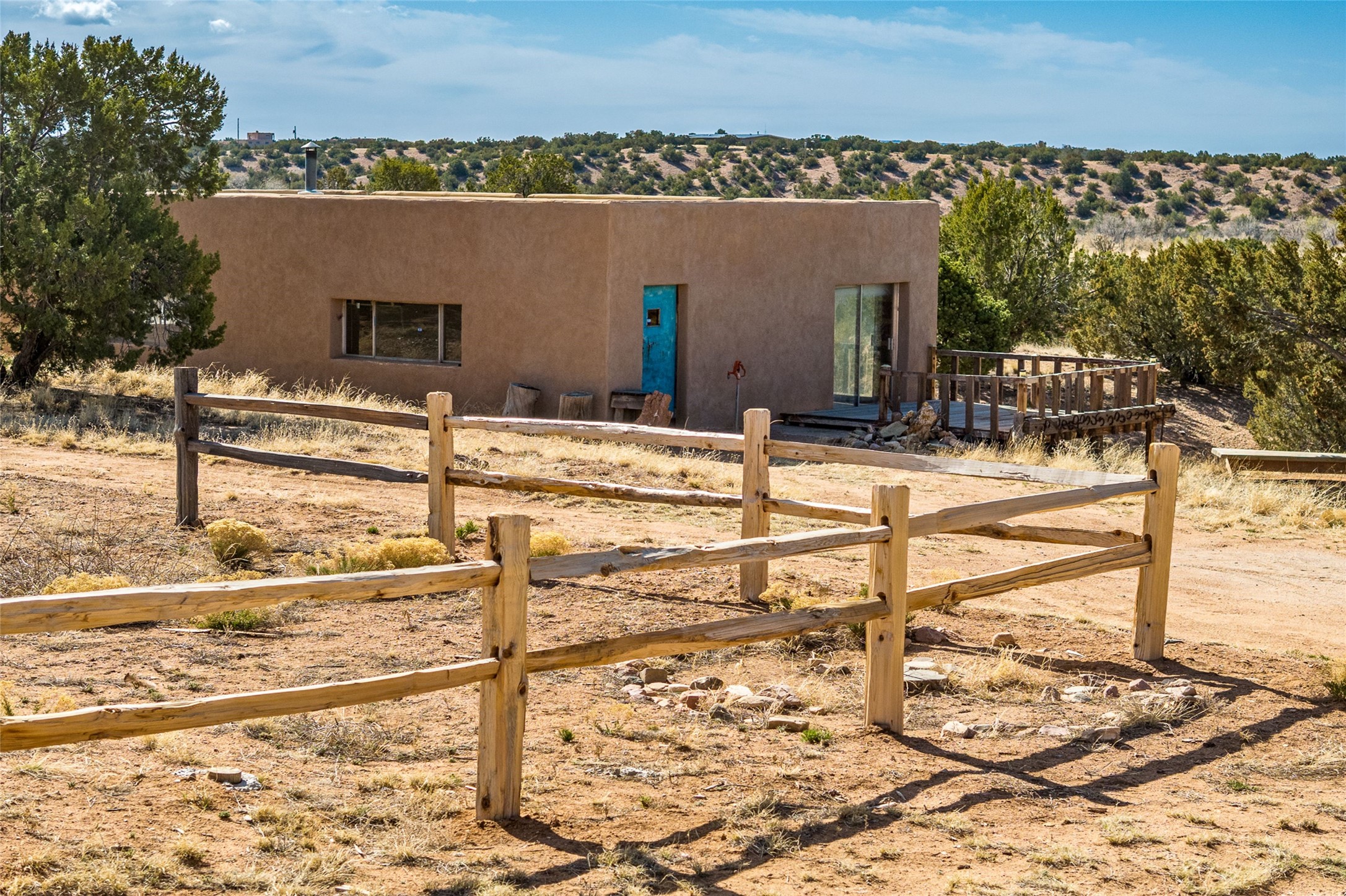 Flat-roofed adobe-style home with a turquoise door, wooden deck, and rustic fence.