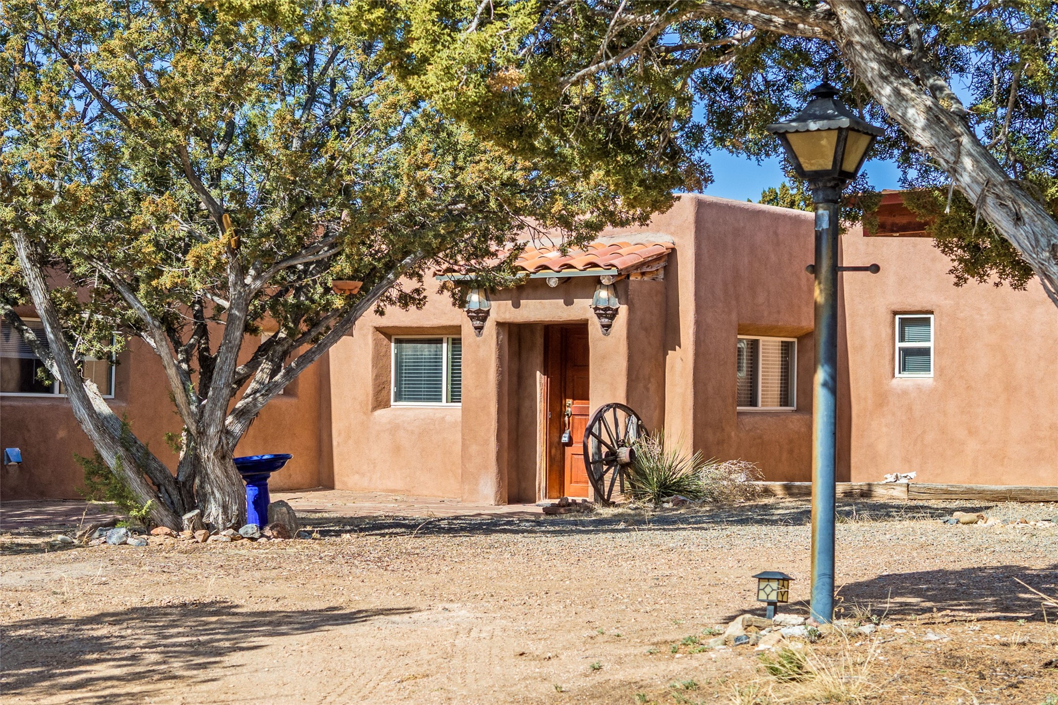 Adobe-style home with a recessed wooden entry, tile awning, mature trees, and gravel yard.