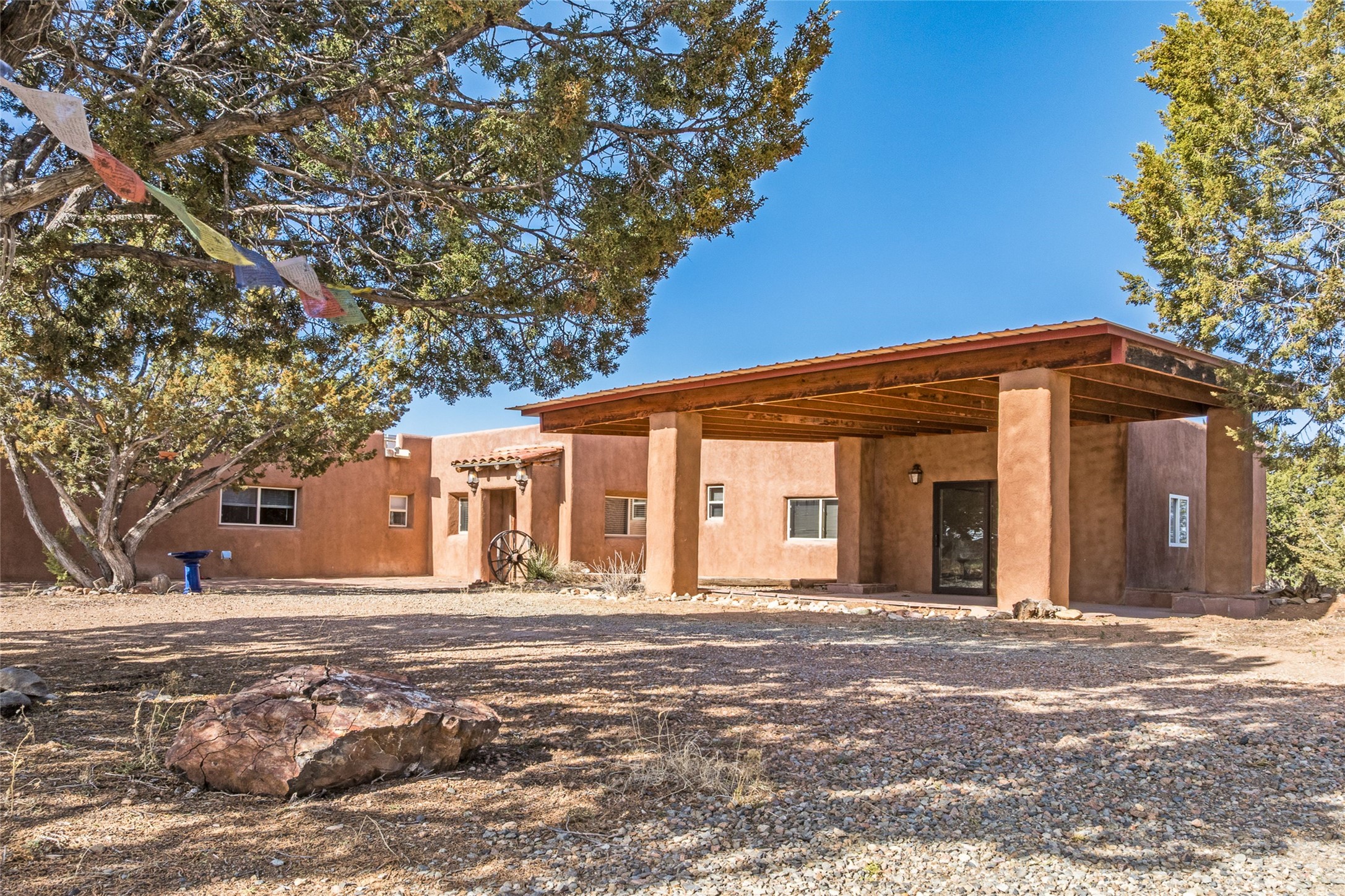 Southwestern stucco home with a broad covered porch, mature trees, and a gravel yard.