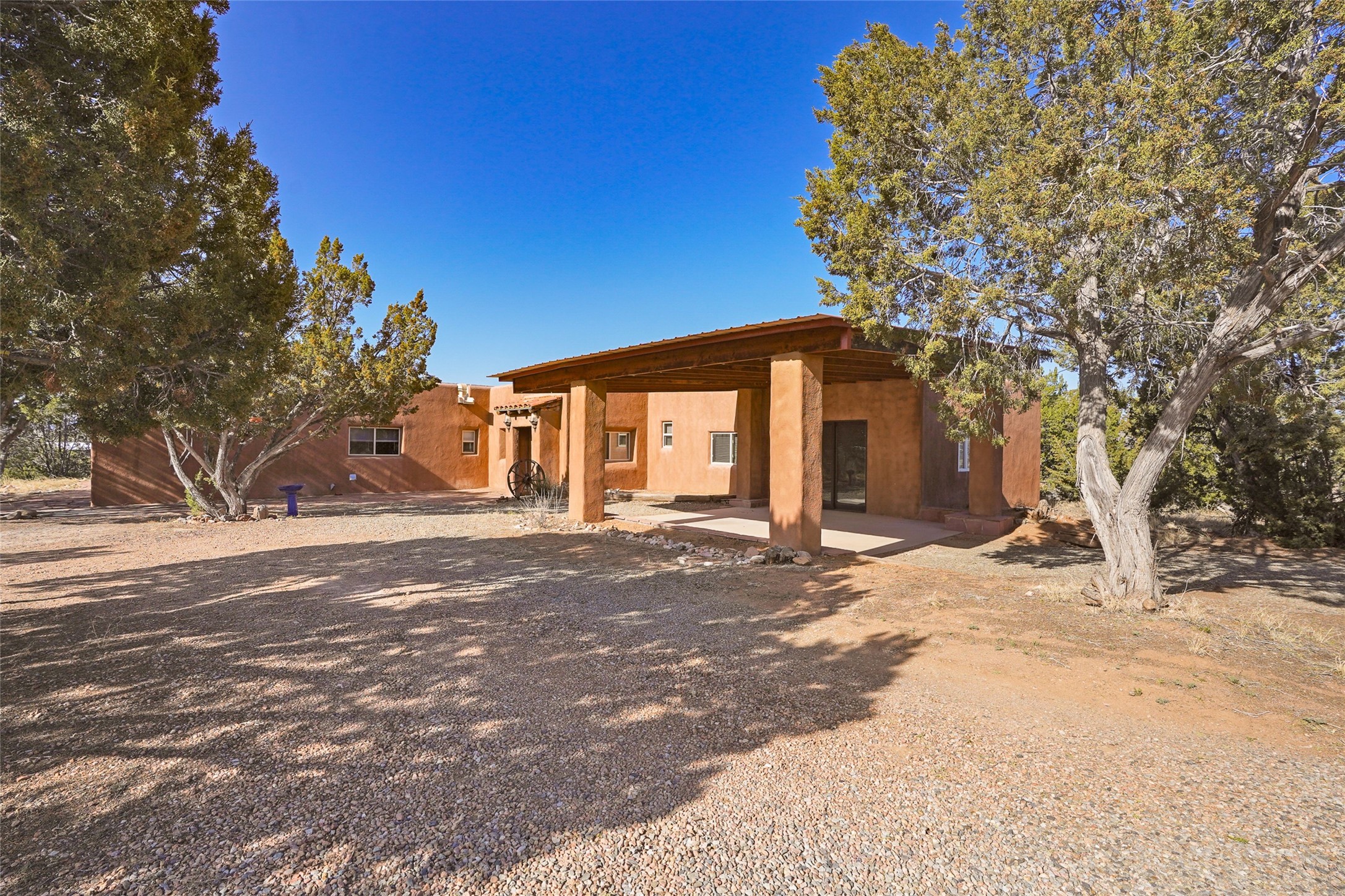 Adobe-style home with a deep covered porch, gravel yard, and mature trees.