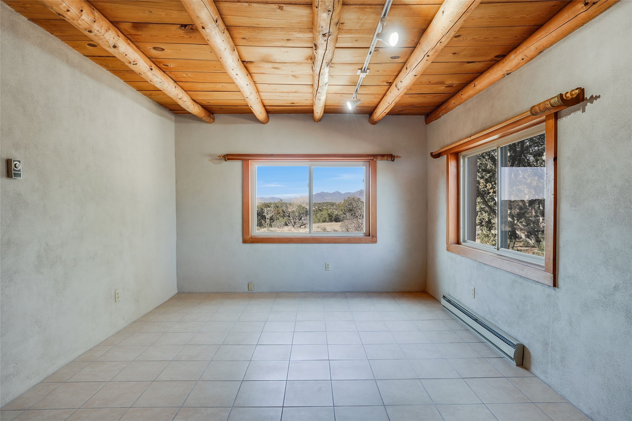 Vacant room with exposed wooden ceiling beams, tiled floor, and mountain views through two windows.