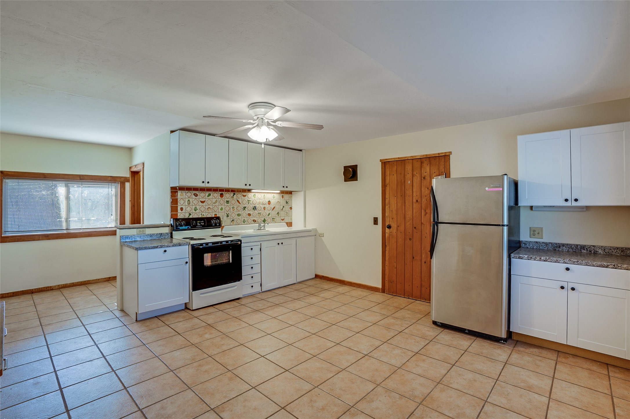 Vacant kitchen with white cabinets, patterned backsplash, tan tile floor, and stainless steel refrigerator.