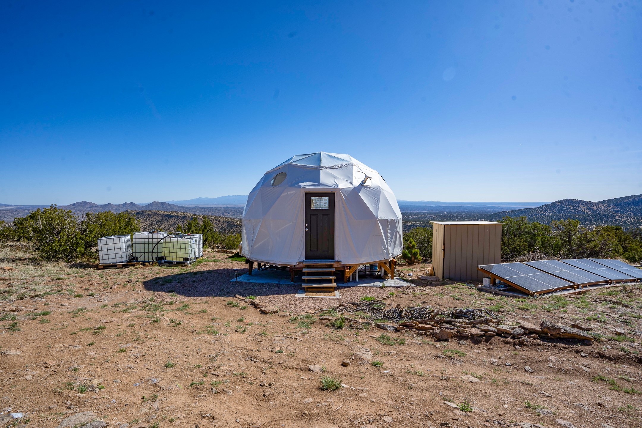 Geodesic dome cabin with solar panels and water tanks on a rugged mountain-view lot.