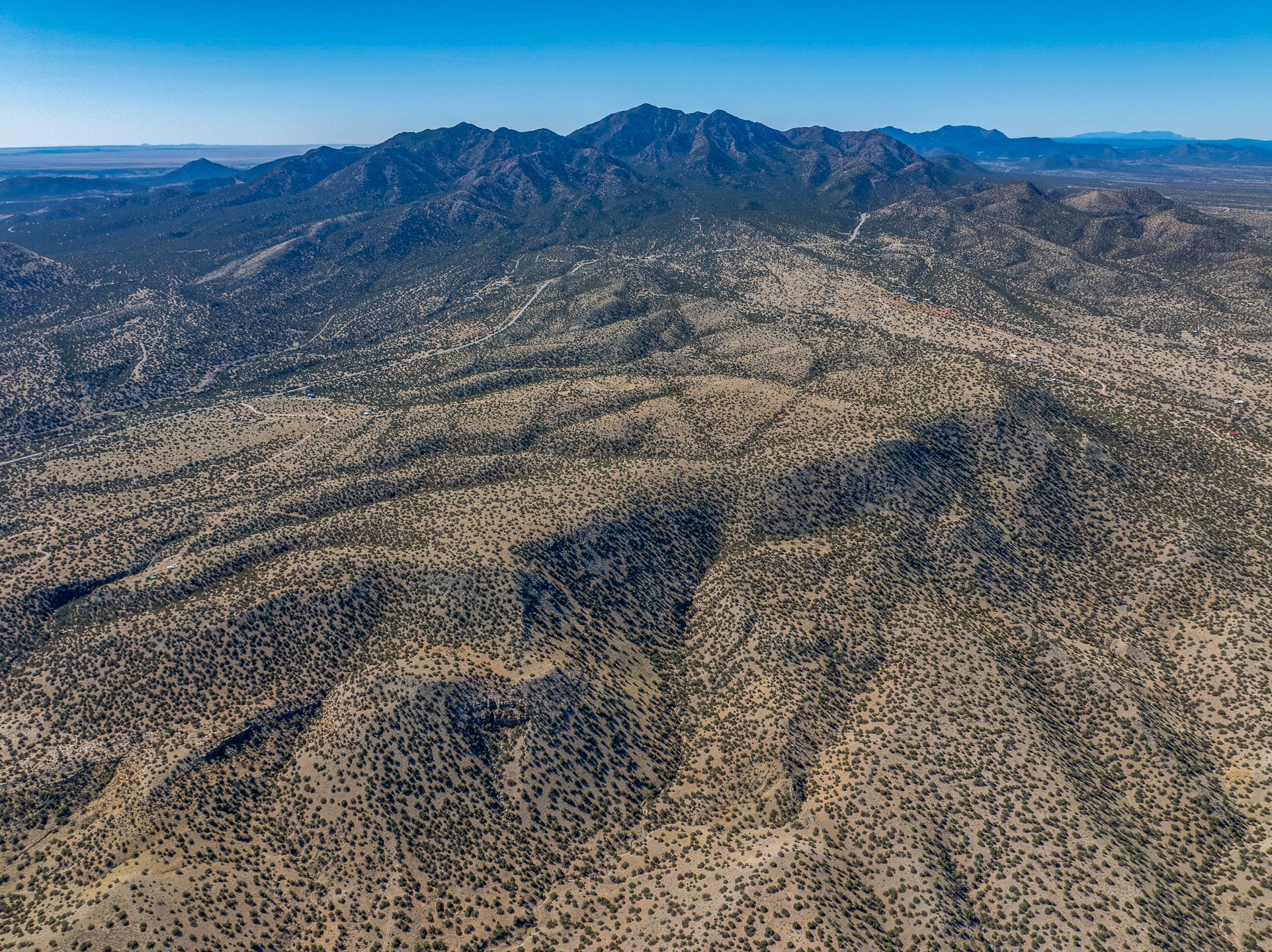 Aerial view of scrub-covered high-desert terrain and dirt roads, with a rugged mountain range beyond.