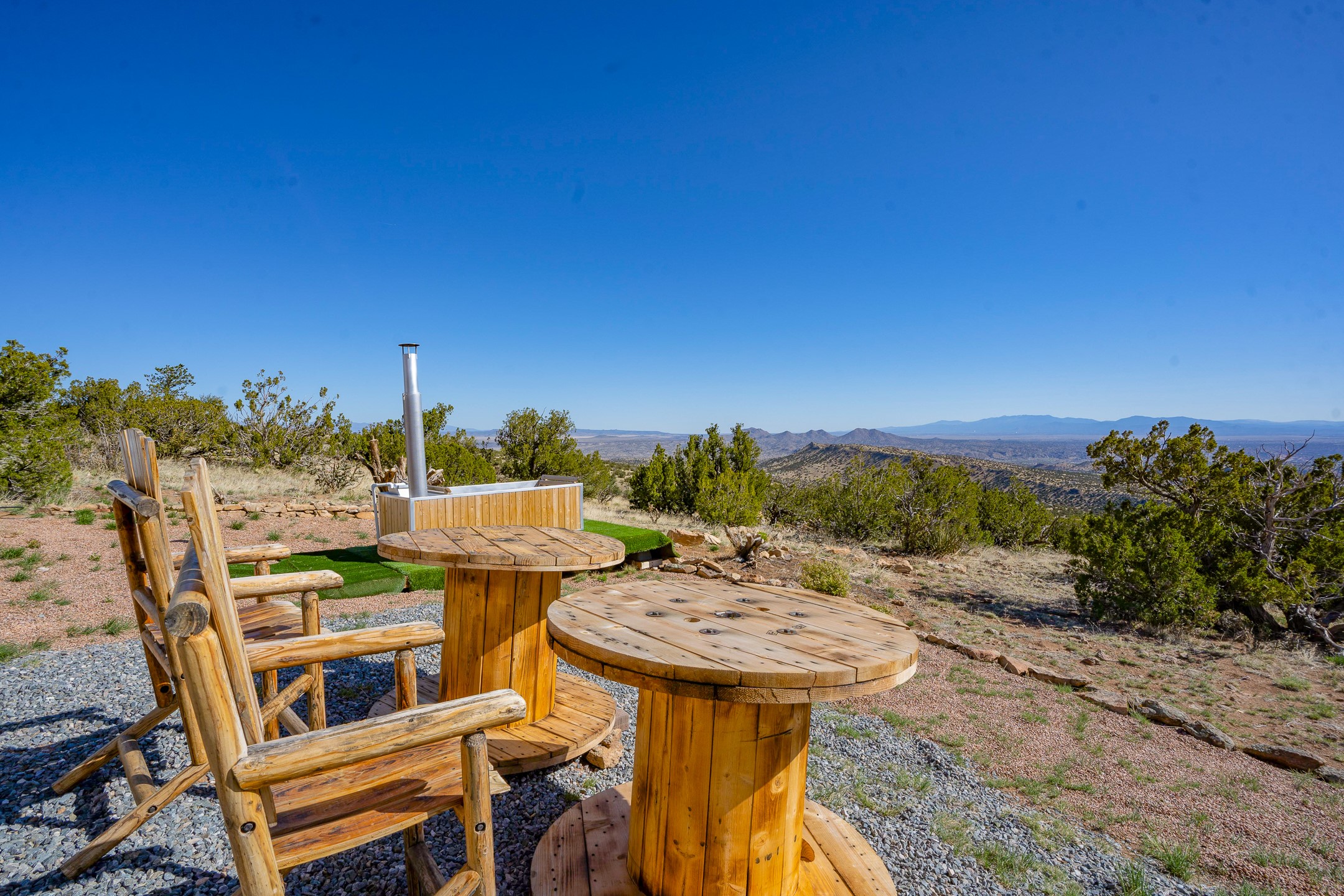 Rustic wooden chairs and spool tables overlook a mountain landscape beside a wood-fired hot tub.
