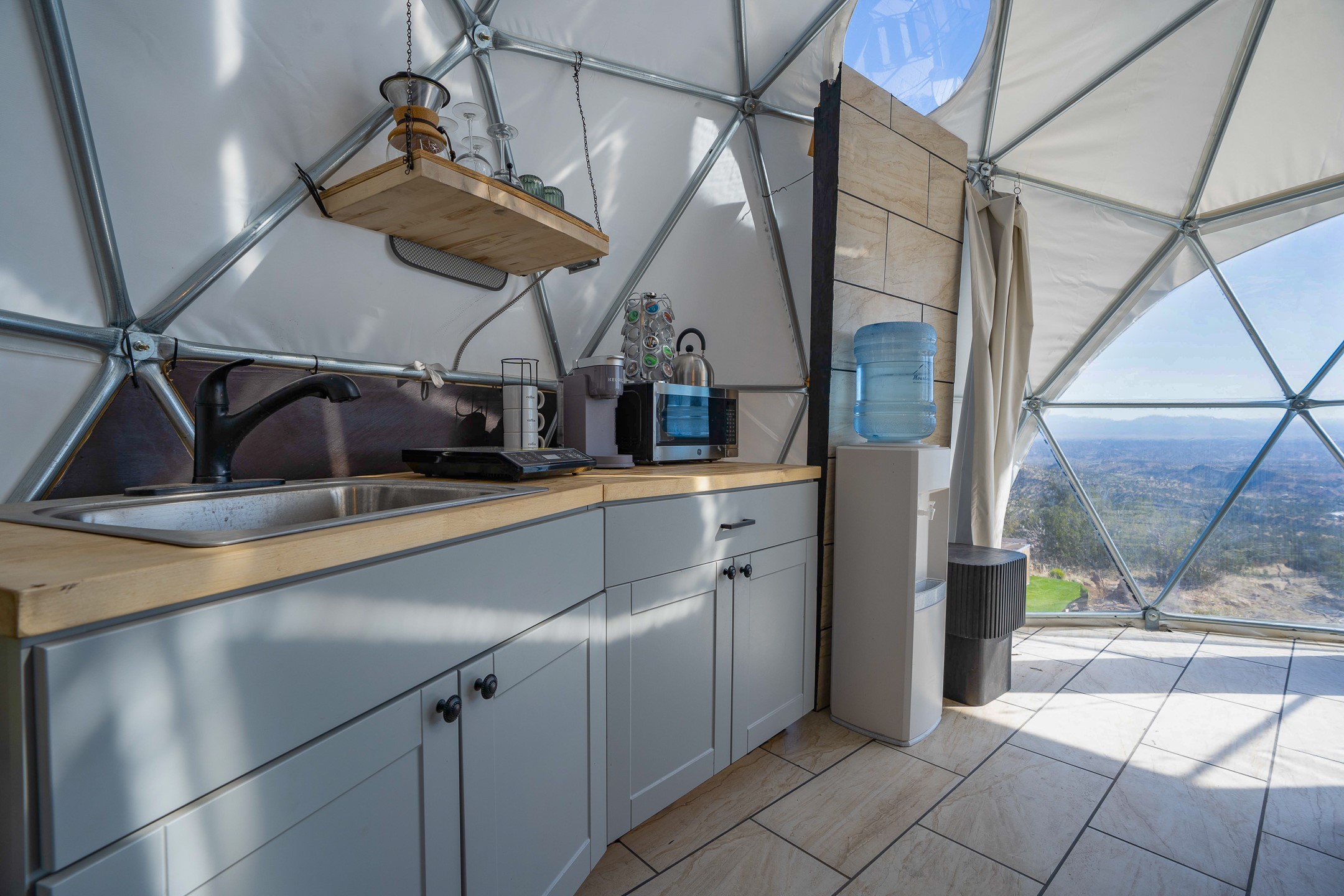 Dome kitchen with gray cabinets and a sink beside panoramic windows overlooking a rugged landscape.