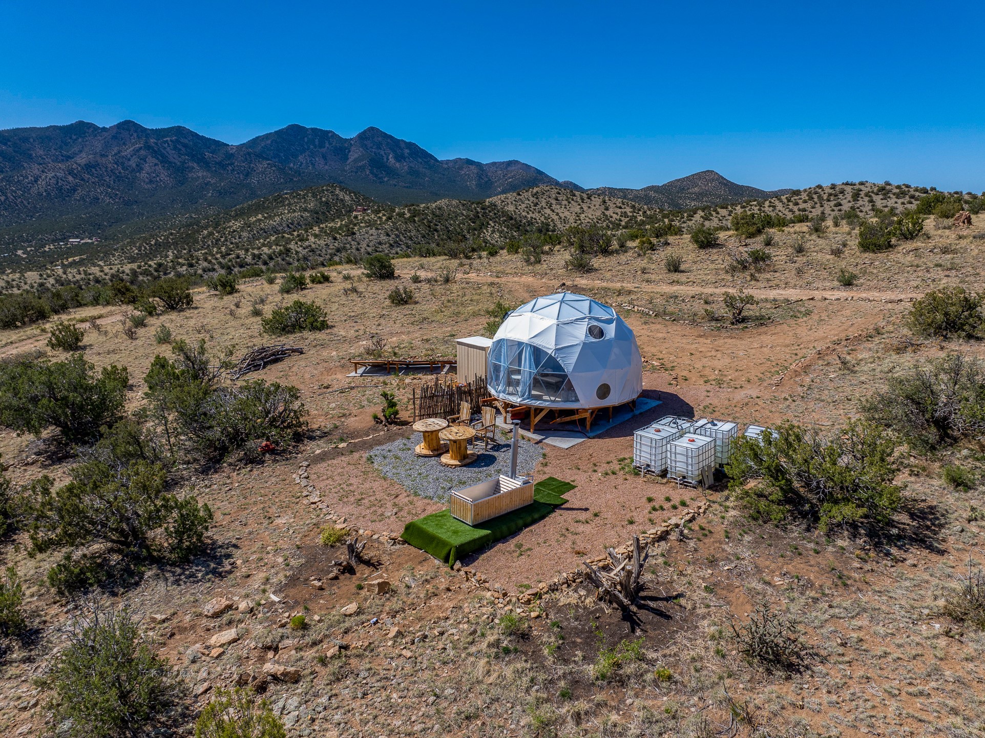 Geodesic dome dwelling on a scrub-covered hillside, with outdoor seating and mountains behind it.