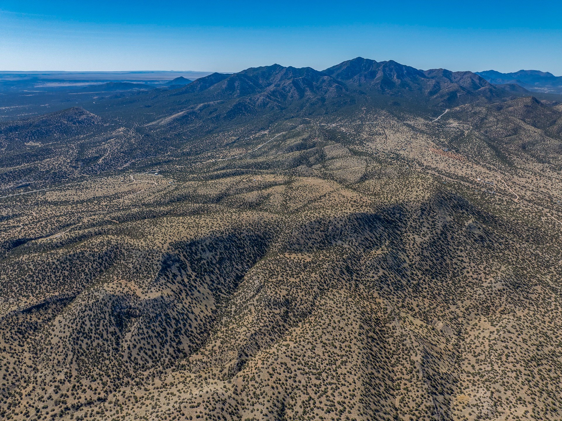 Aerial view of scrub-covered hills and rugged mountains beneath a clear blue sky.