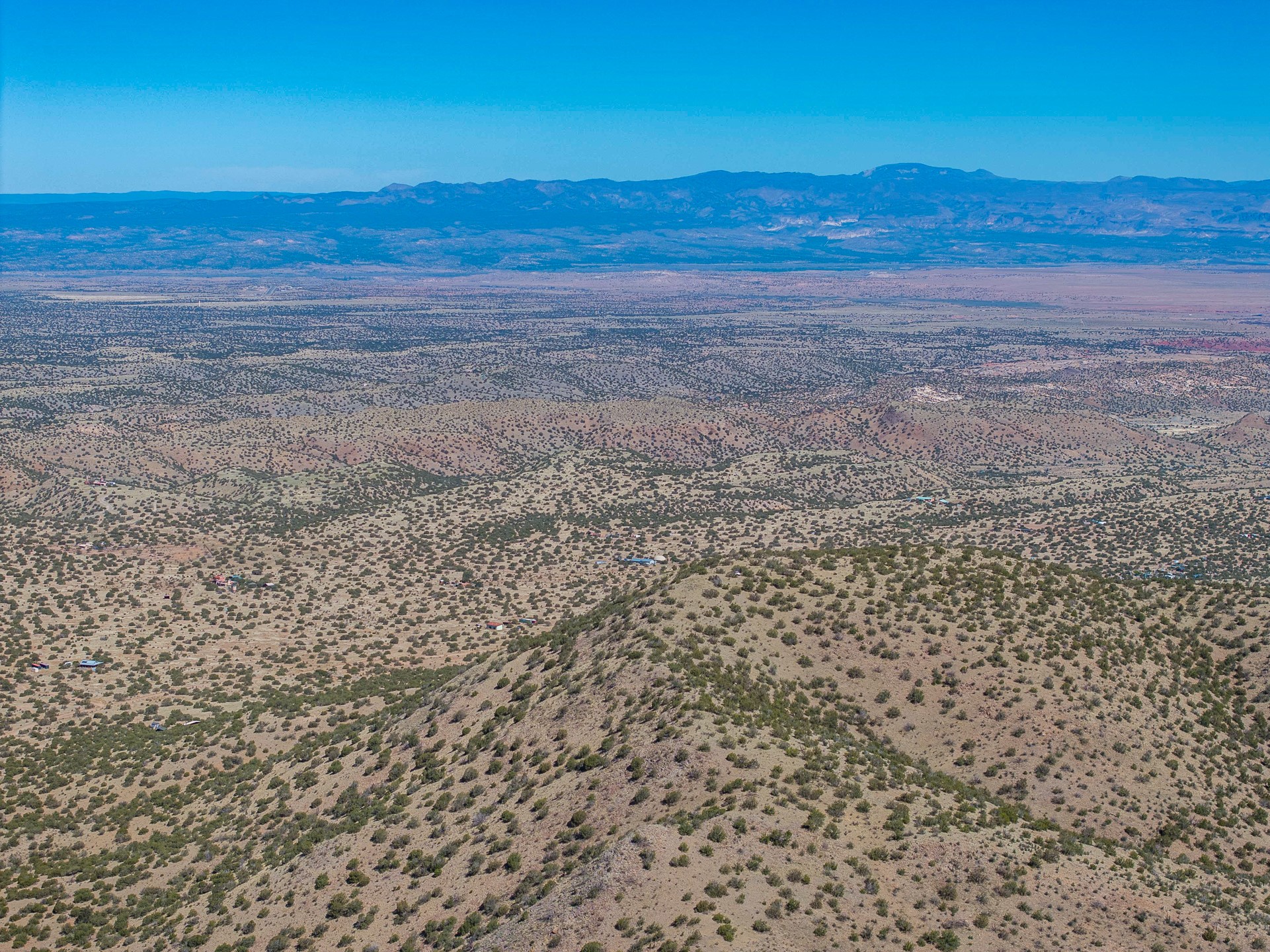 Aerial view of scrub-covered desert hills and scattered buildings, with distant mountain ranges.