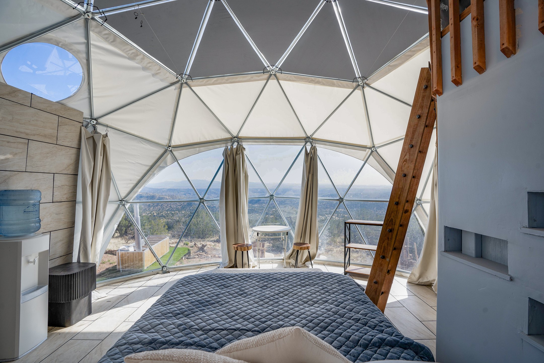 Dome bedroom with a quilted bed, timber ladder, and panoramic views through triangular windows.