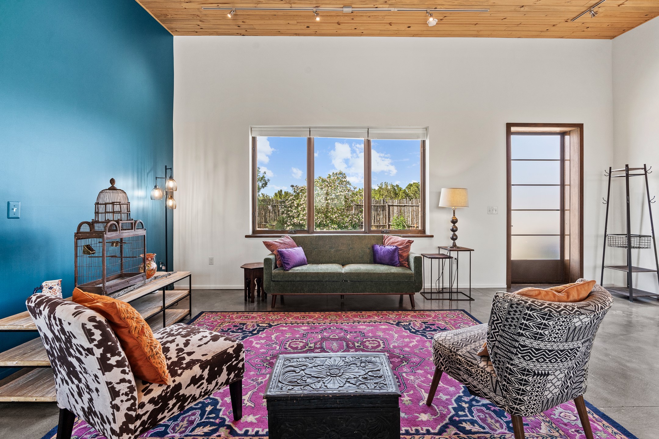 Furnished living room with a teal accent wall, wood ceiling, patterned chairs, and magenta rug.
