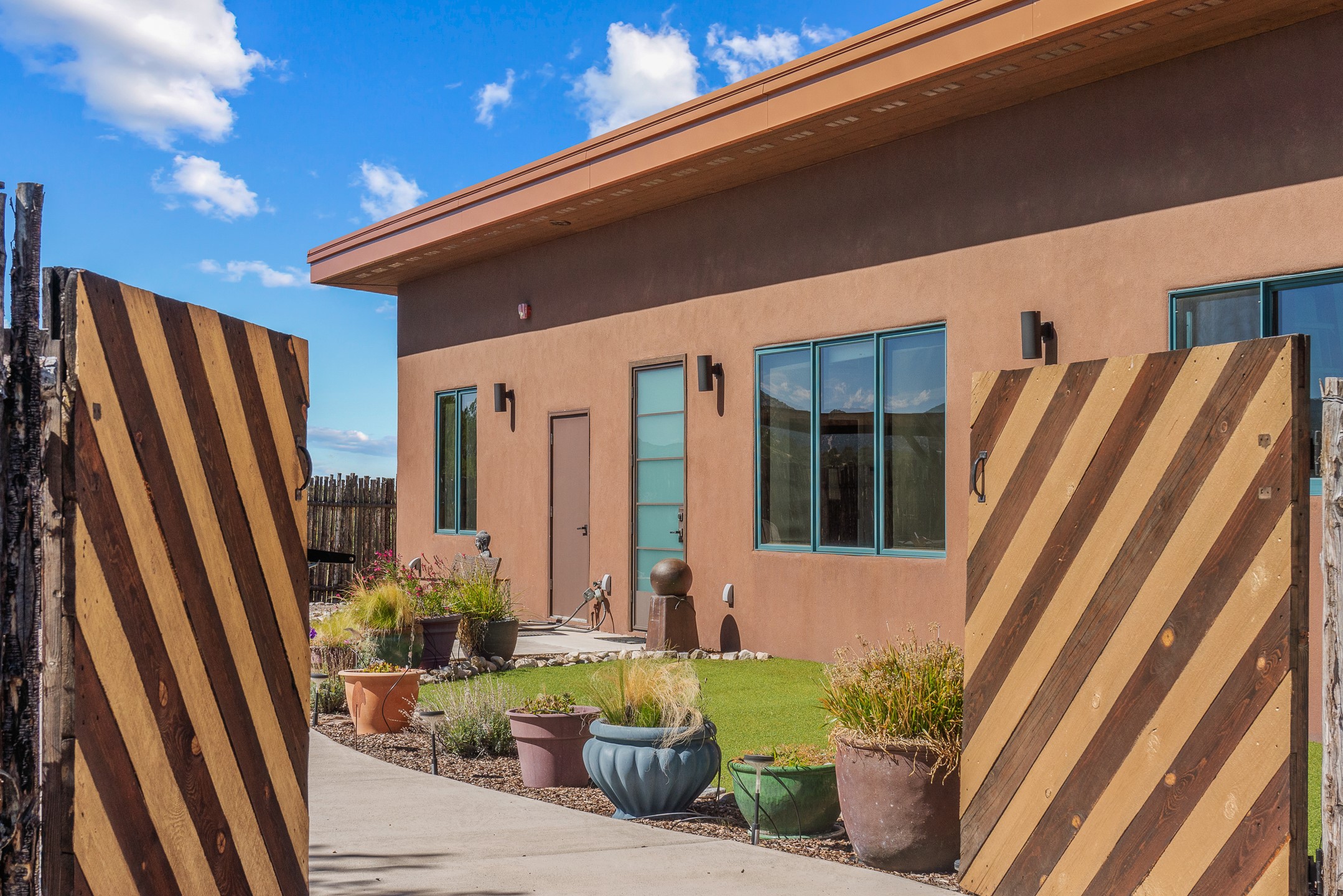 Modern tan stucco home behind open diagonal-plank gates, with patio planters and large windows.