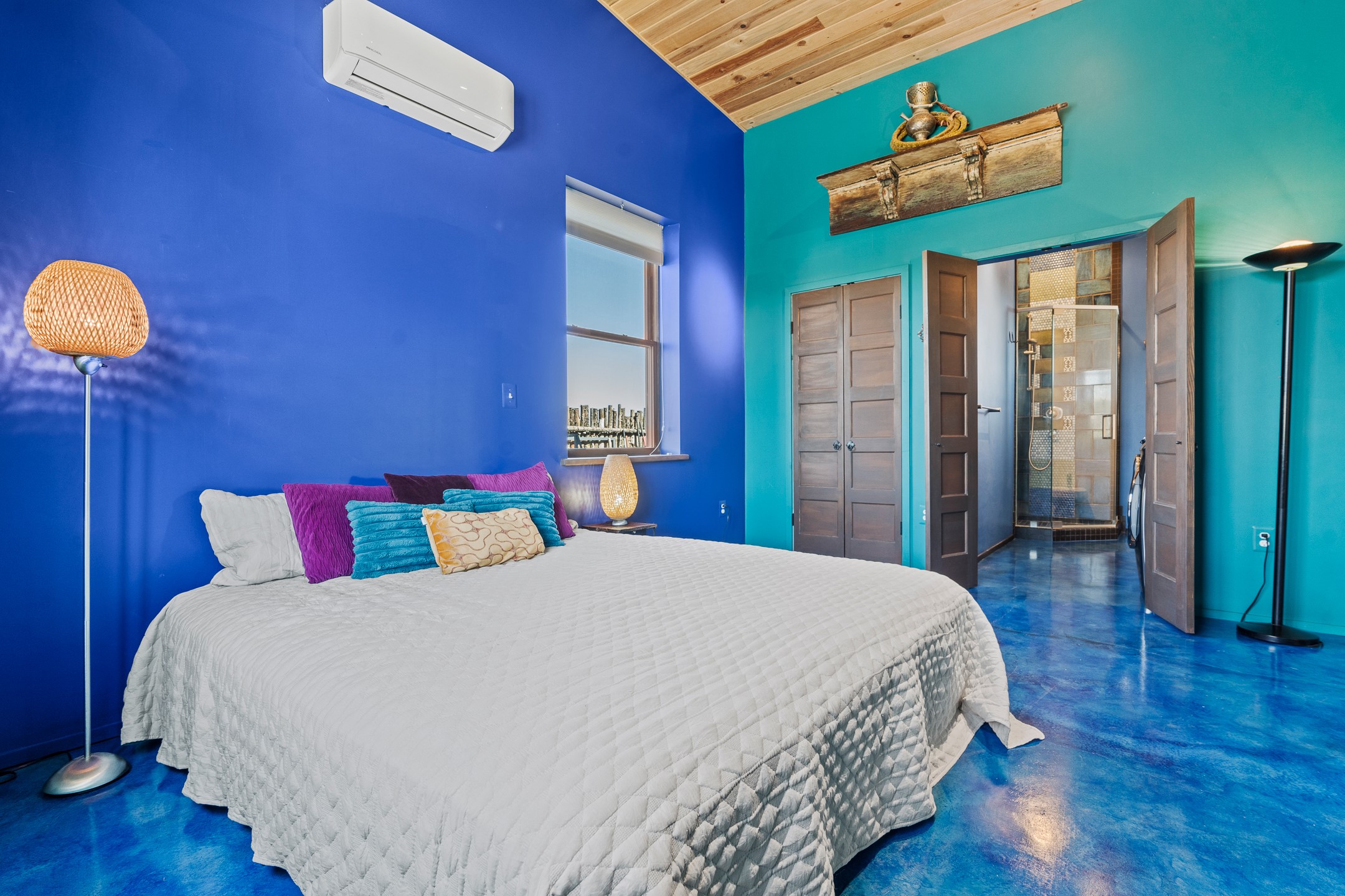 Bedroom with a white bed, vivid blue walls, polished blue floor, and an open doorway to a tiled shower.