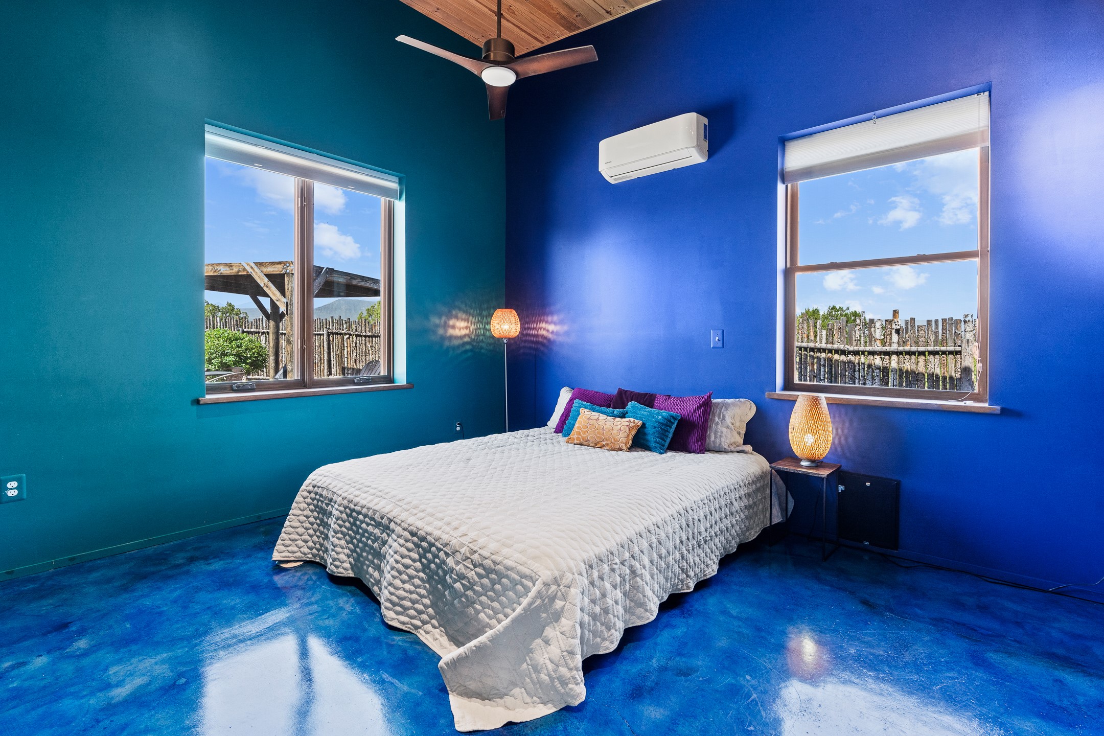 Bedroom with vivid teal and blue walls, a white quilted bed, and windows overlooking a fenced yard.