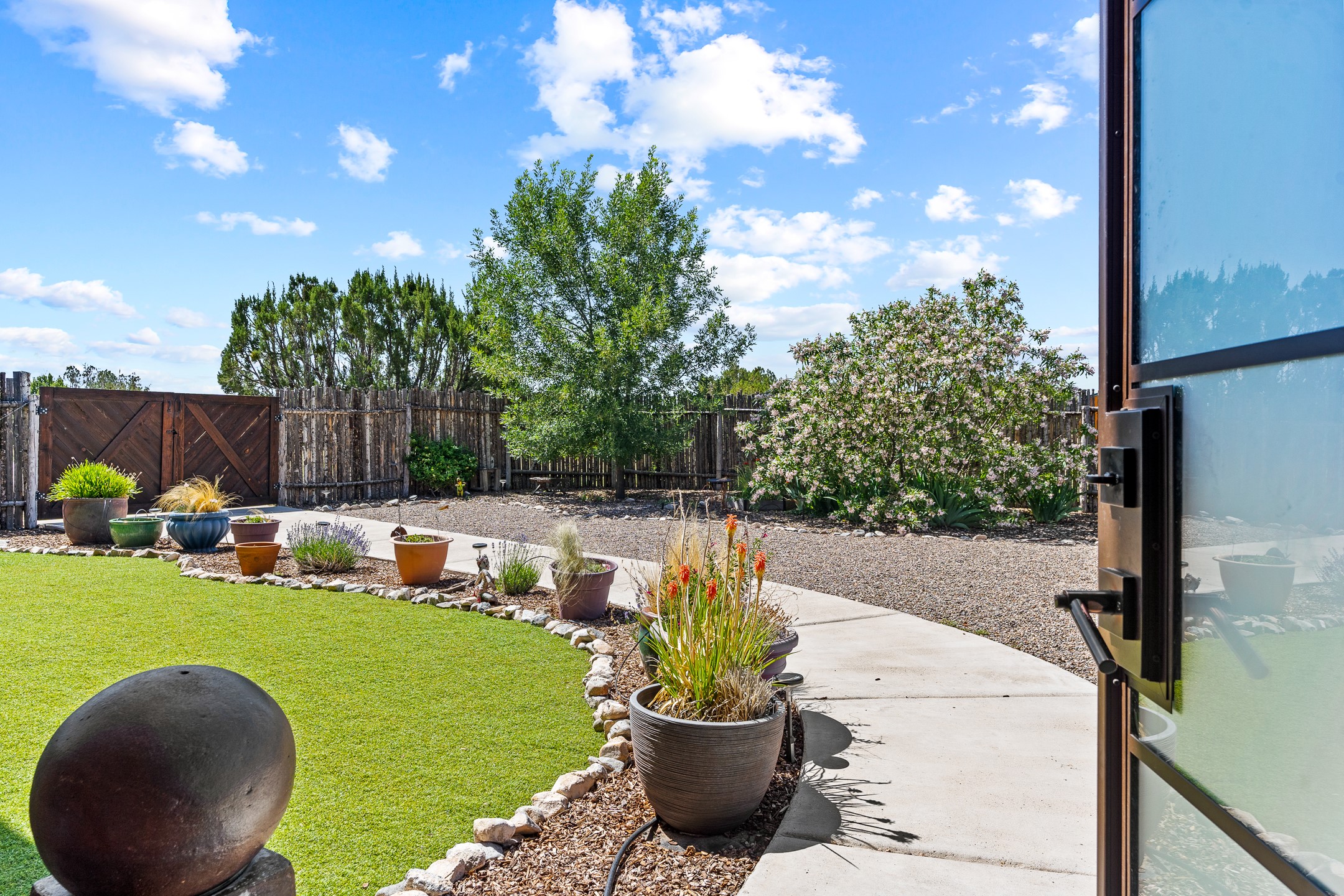 Landscaped backyard with a lawn, potted plants, flowering shrubs, and a curved walkway beside an open glass door.