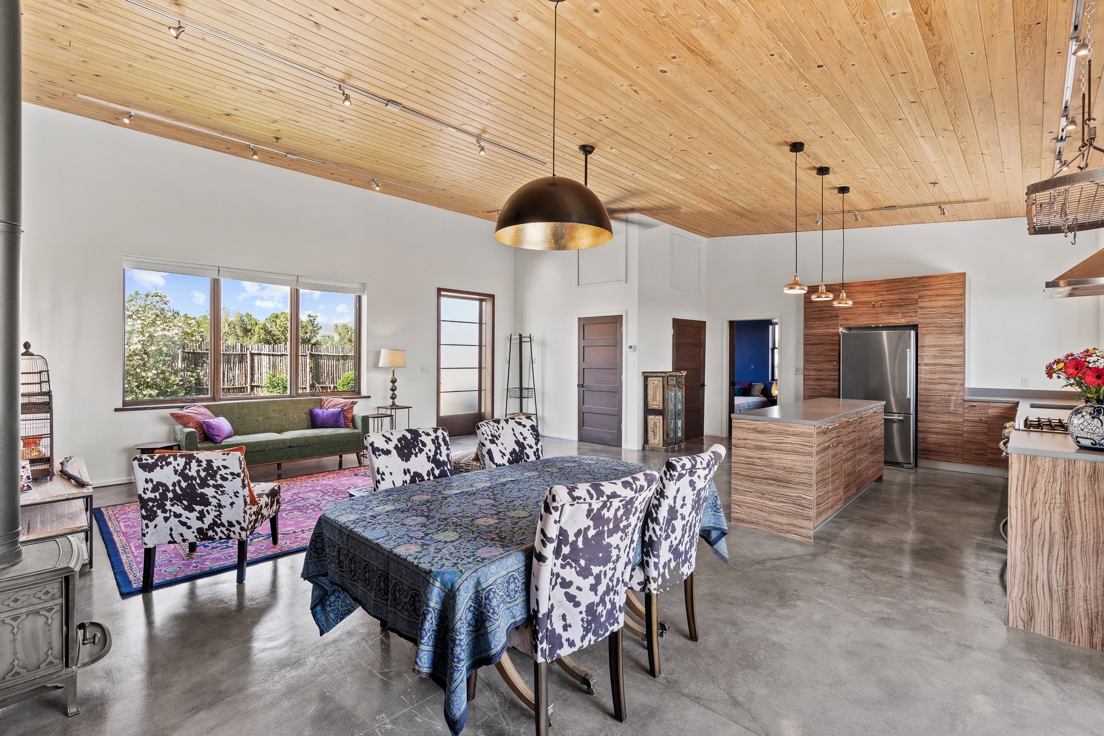 Open-plan kitchen and dining area with wood ceiling, paneled island, patterned chairs, and large yard-facing windows.