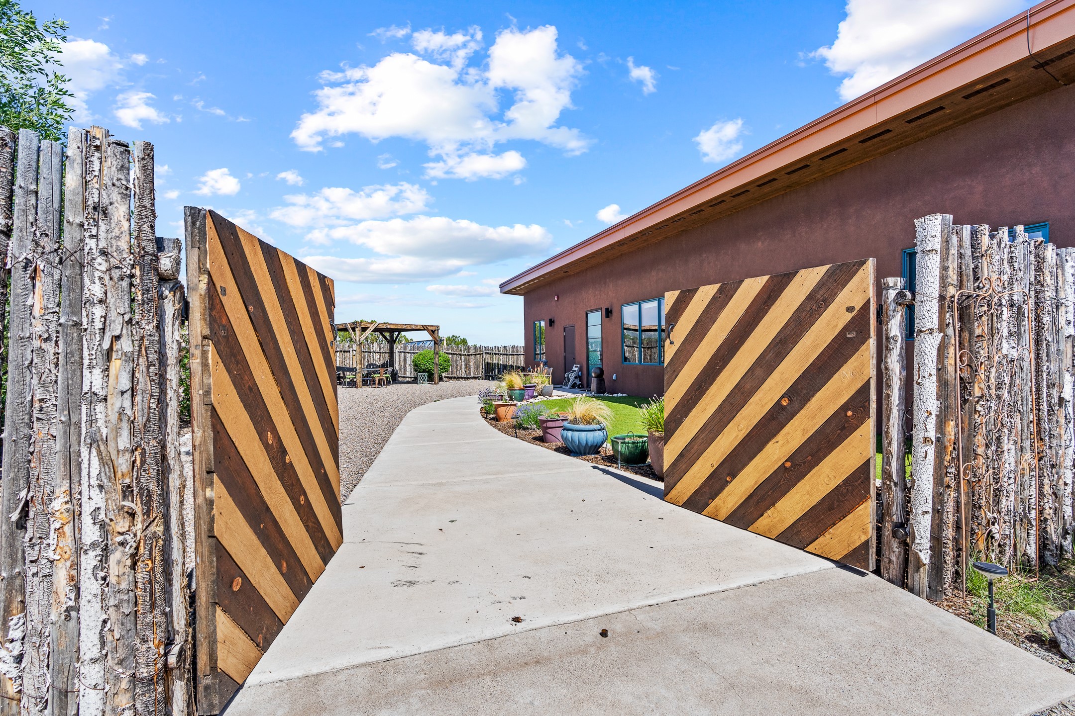 Home entrance framed by open striped wooden gates, with a concrete drive and colorful planters.