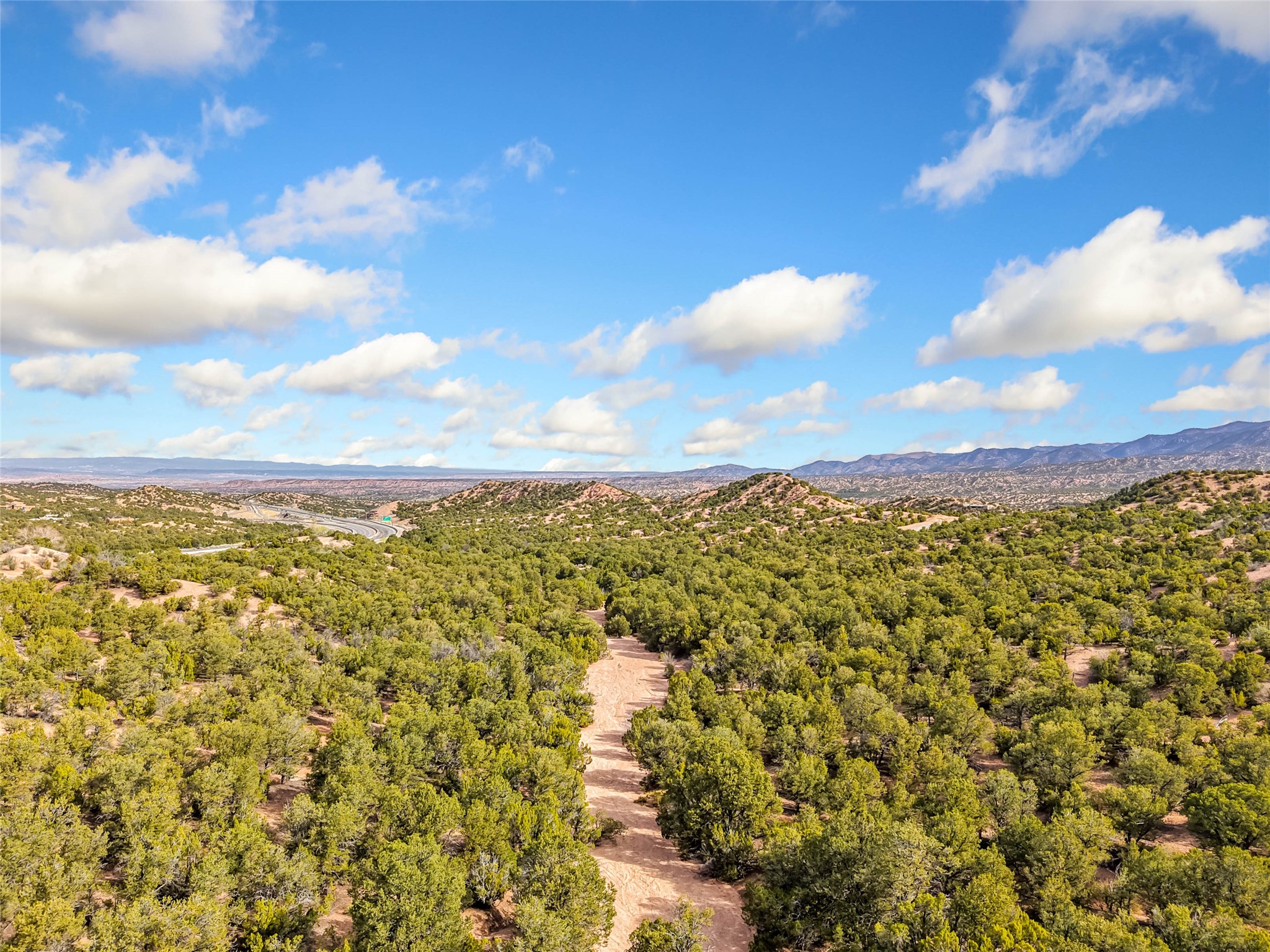 Aerial view of scrub-covered land with a dirt track, distant mountains, and a winding road.