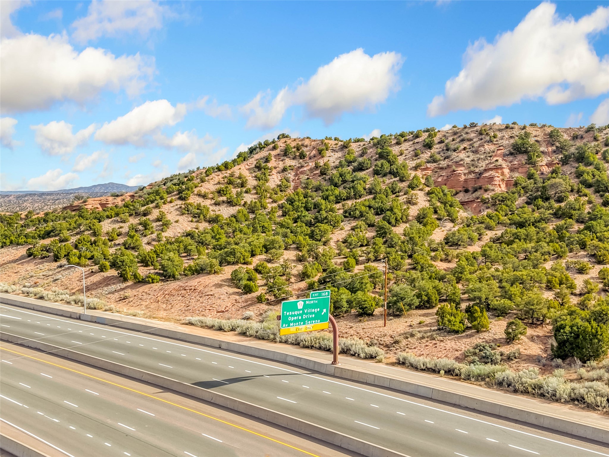 Rugged, juniper-covered land with red rock outcrops rises behind a highway and exit sign.
