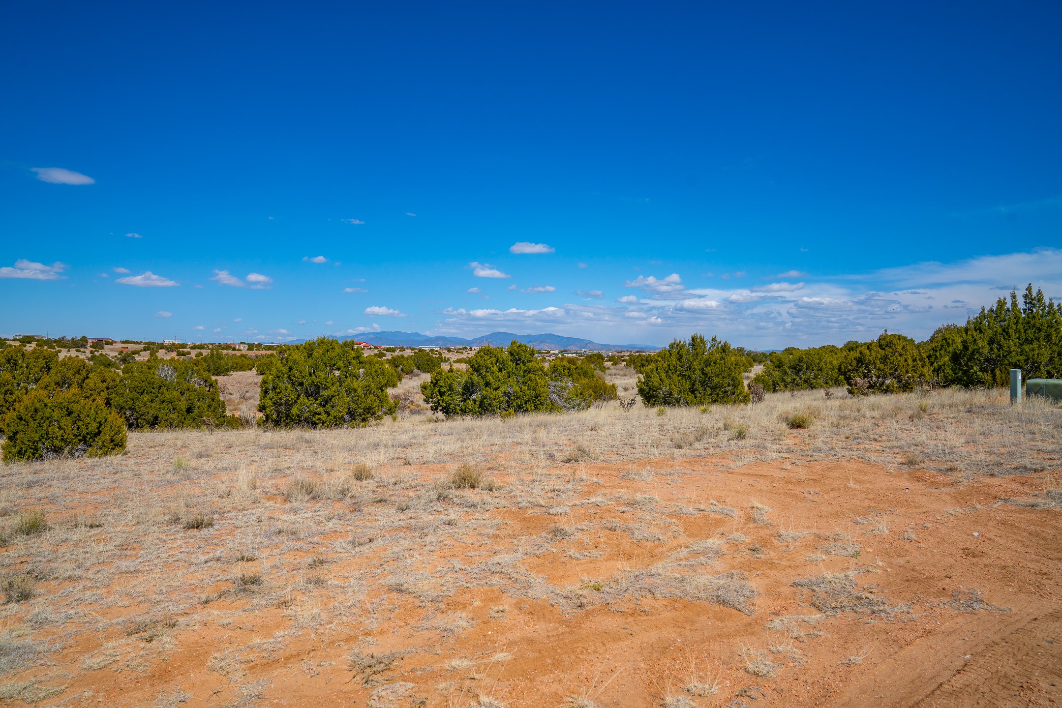 Vacant reddish-soil lot with juniper shrubs and distant mountains beneath a wide blue sky.