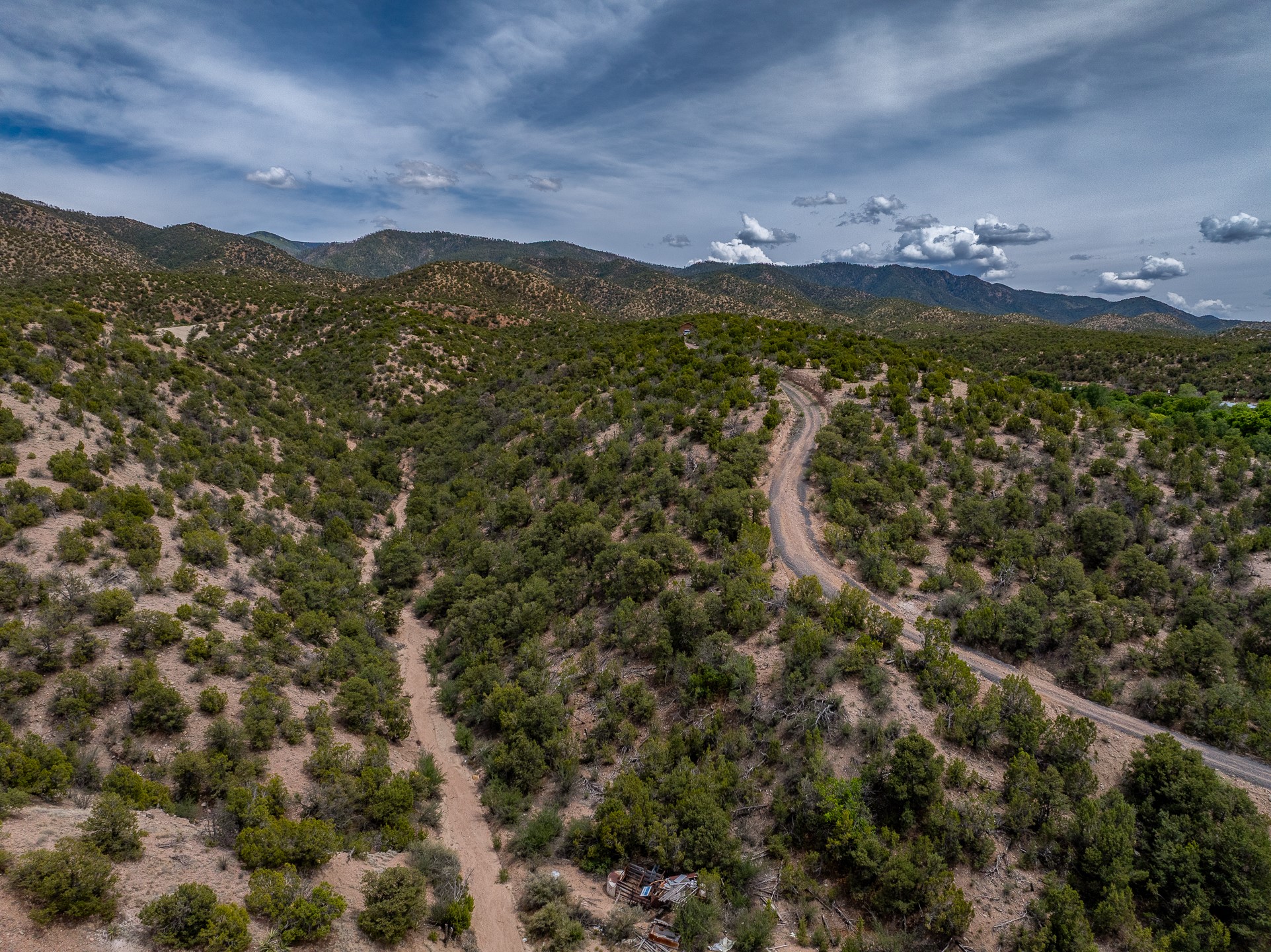 Aerial view of rugged, shrub-covered land with a winding dirt road and mountain ridges beyond.