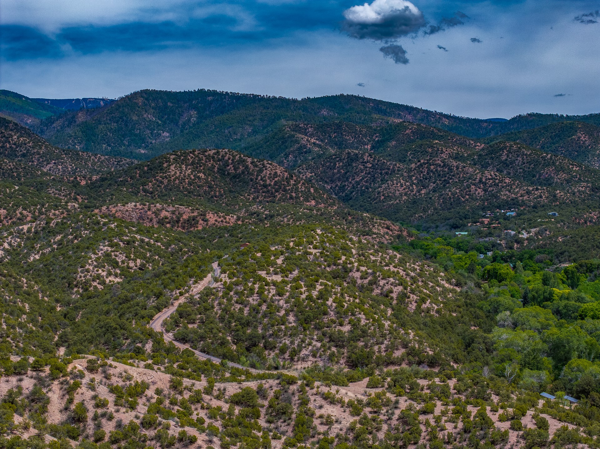 Aerial view of rugged hills, a winding road, distant mountain ridges, and scattered homes in a green valley.