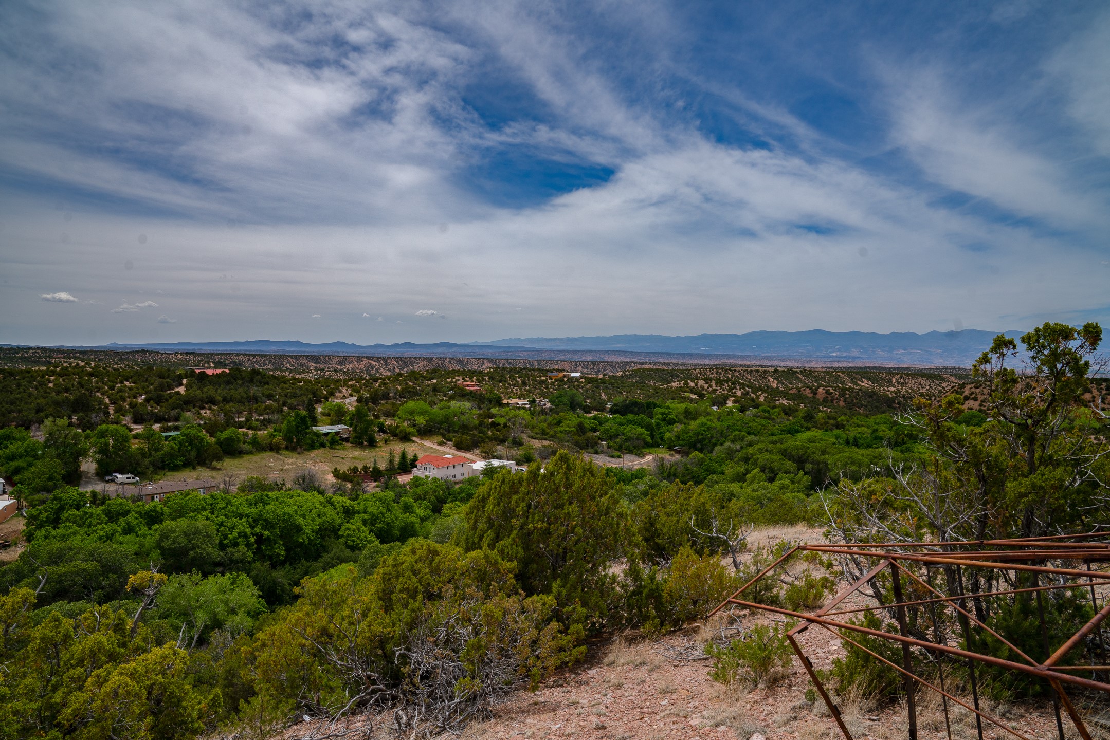 Panoramic mountain view over wooded terrain, with a rocky path and rust-colored railing in the foreground.
