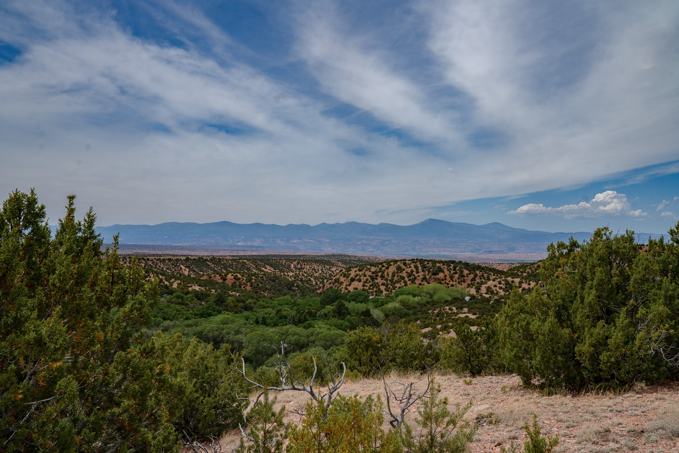 High-desert hills and juniper trees overlook a distant mountain range beneath a sweeping, cloud-streaked sky.