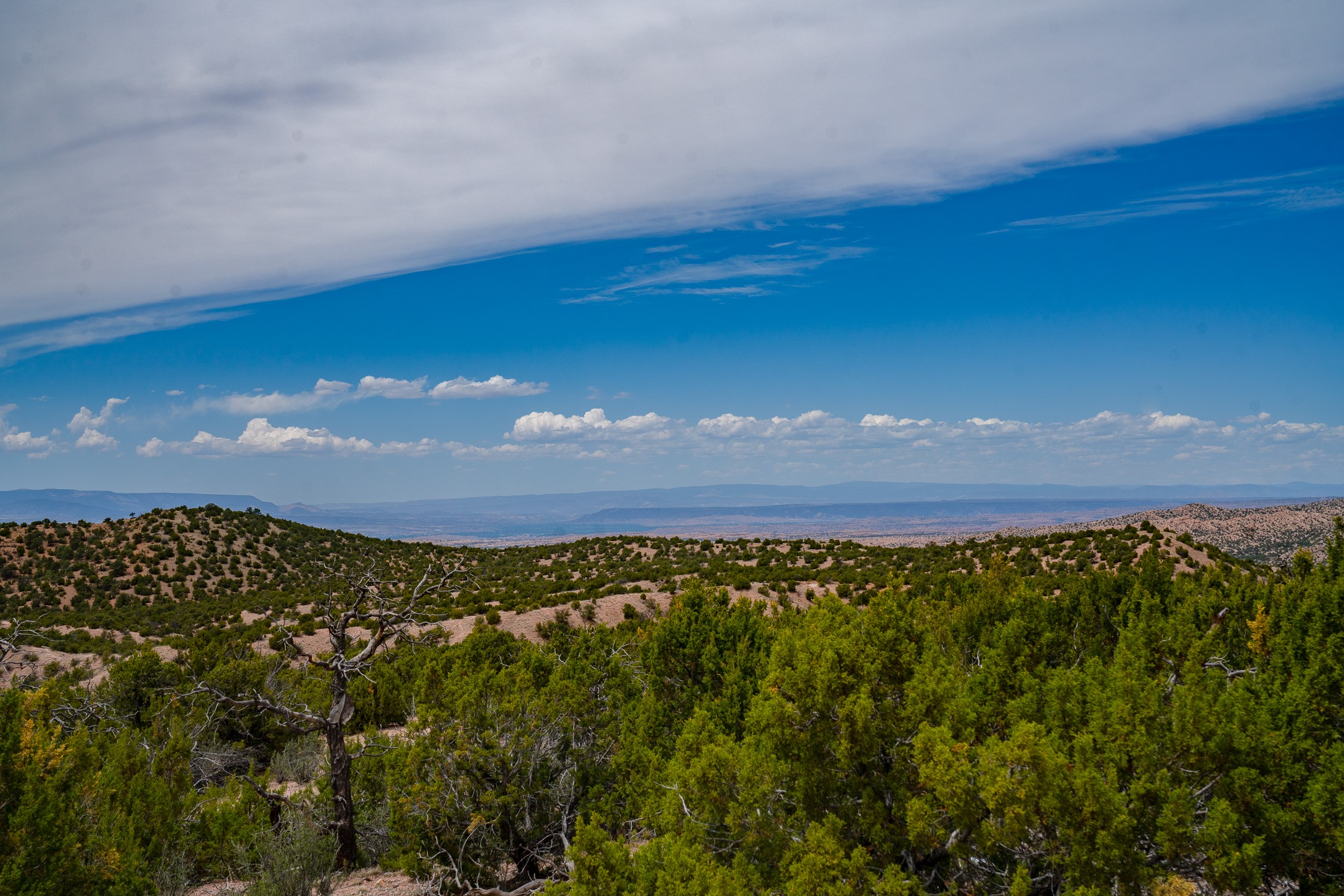 Panoramic mountain view beyond scrub-covered hills and green shrubs beneath a cloud-filled blue sky.