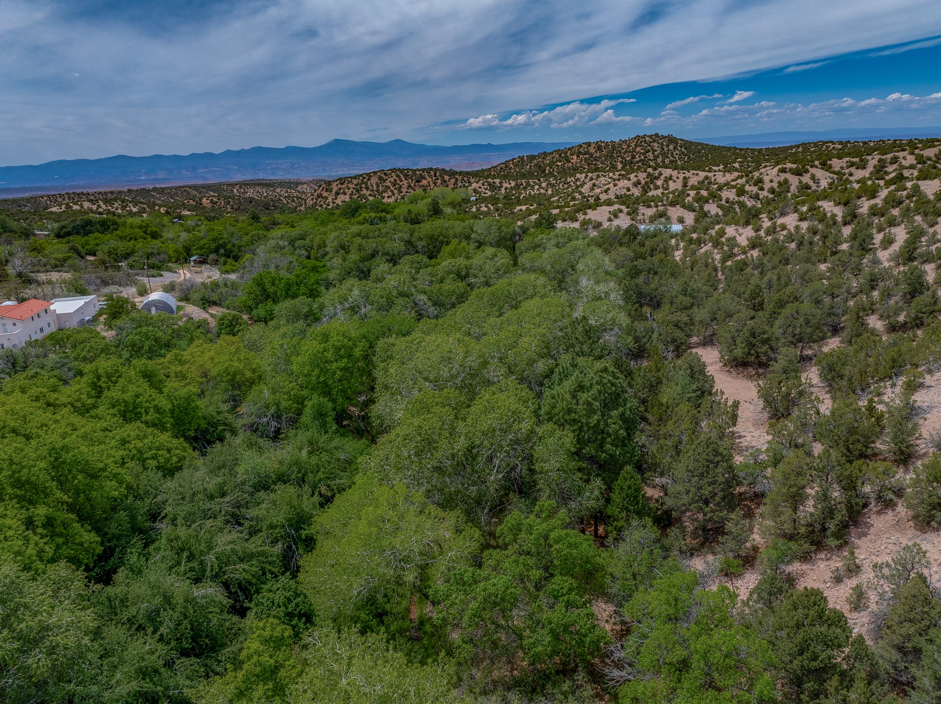 Aerial view of wooded hills and scattered buildings, with distant mountains and a dry slope.