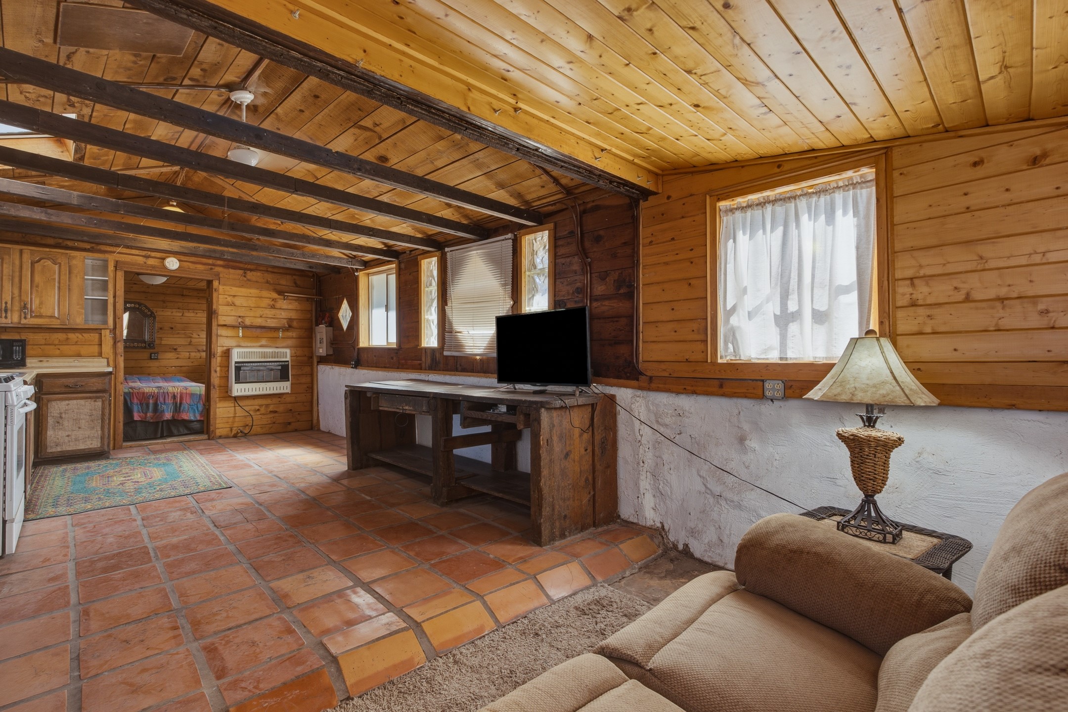 Rustic living room with exposed beams, terracotta tile flooring, and a bedroom visible through the doorway.