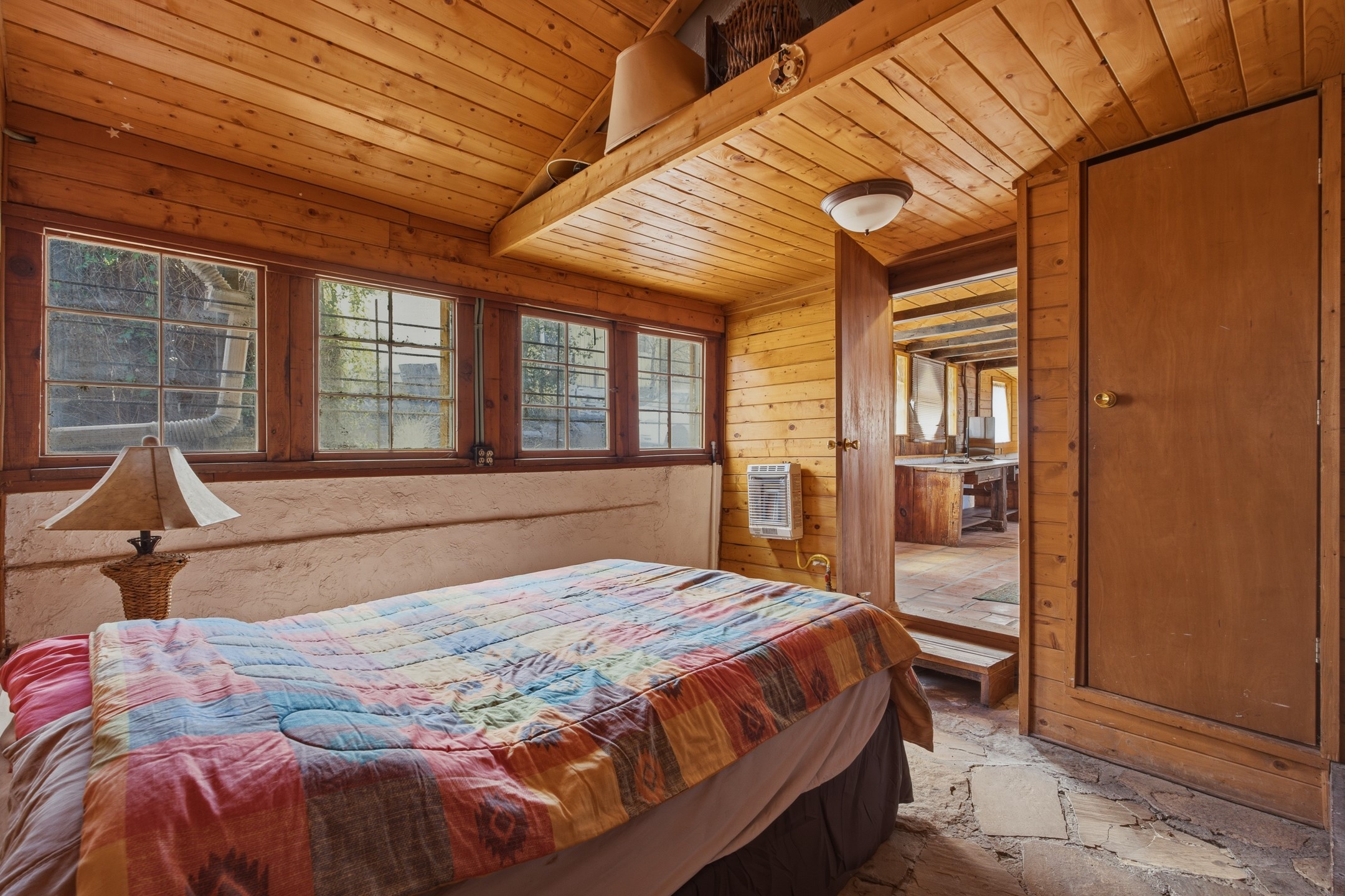 Bedroom with a patchwork-quilt bed, wood-plank ceiling, multiple windows, and a doorway to the kitchen.