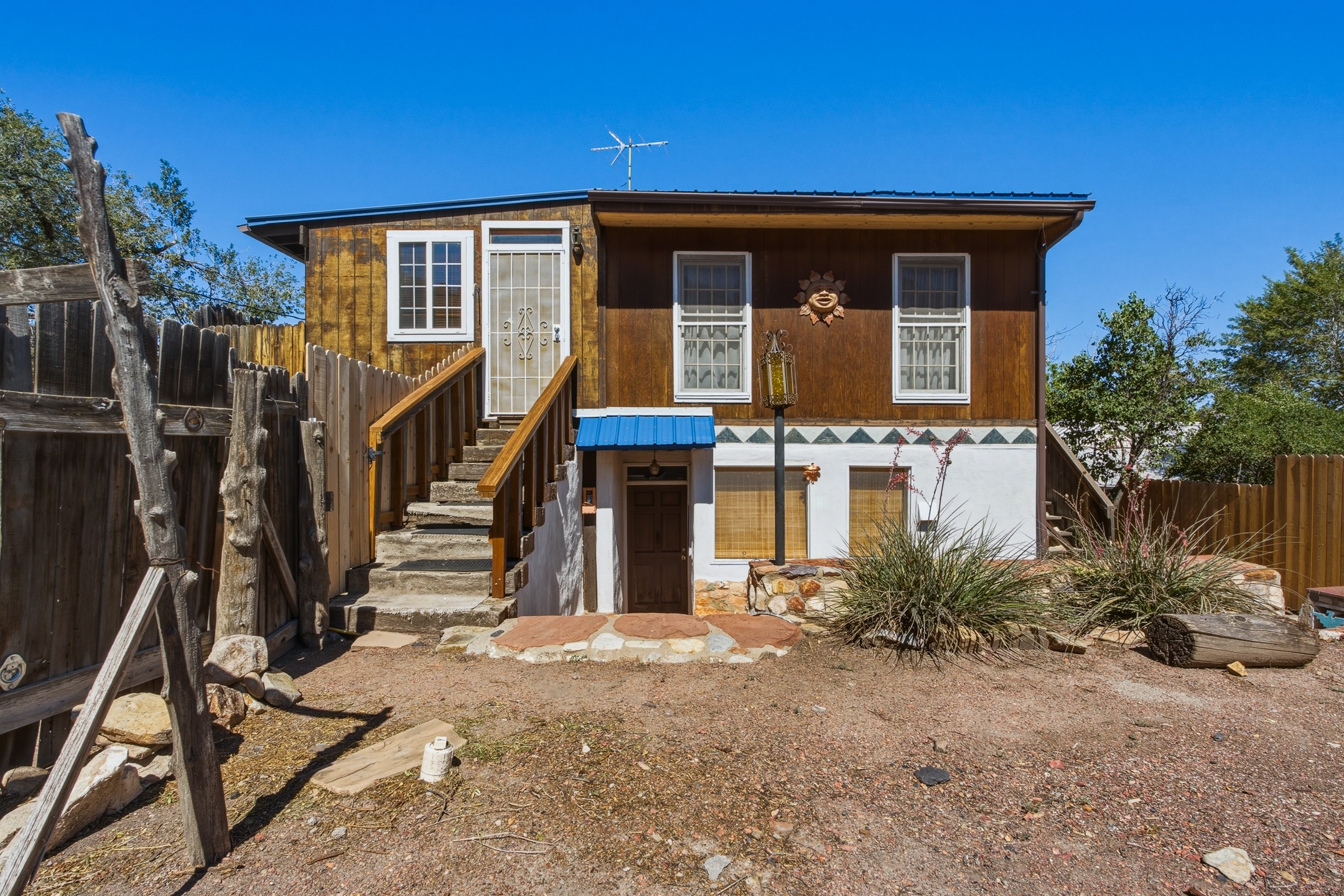 Two-level brown and white home with a blue awning, outdoor staircase, and fenced gravel yard.