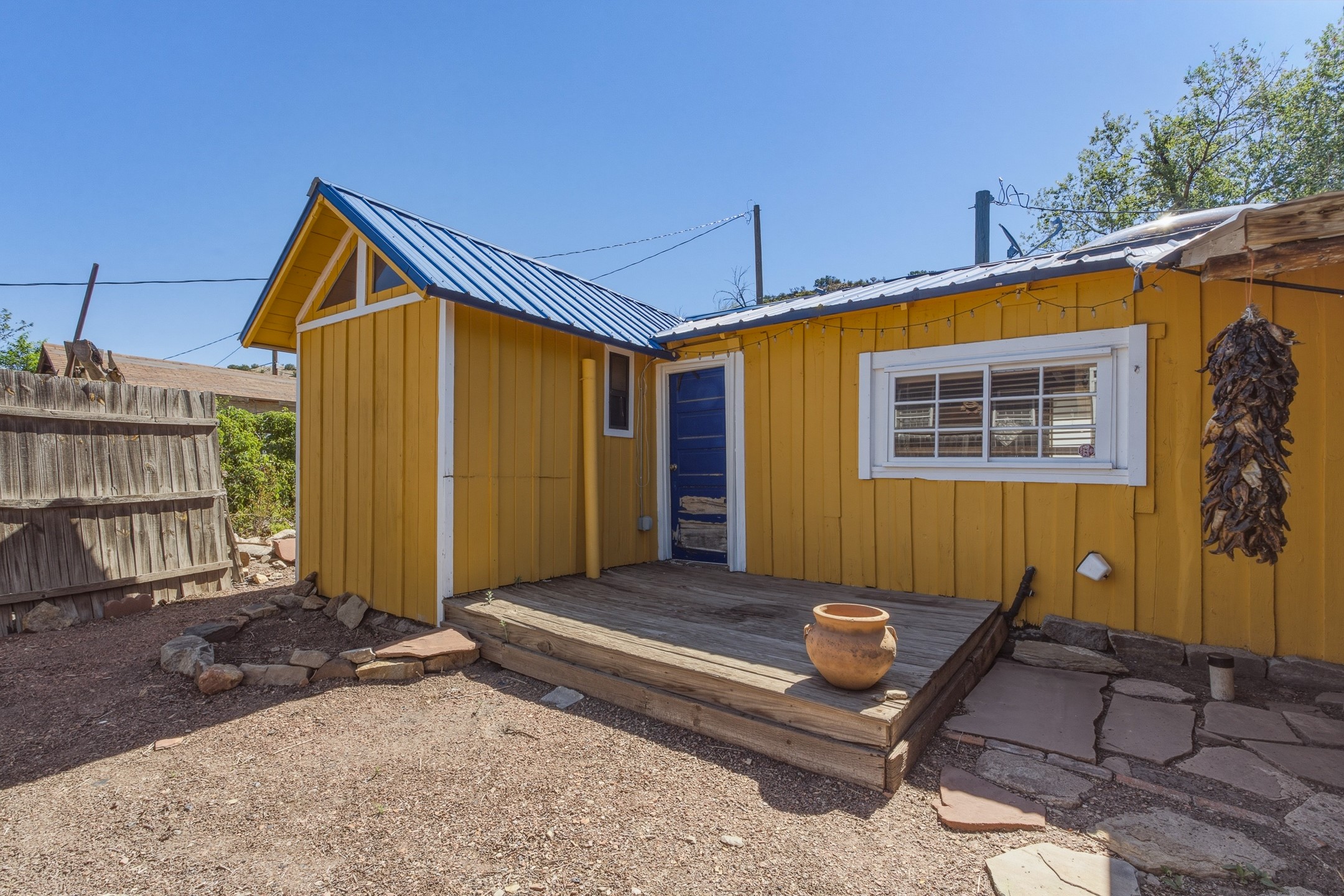 Mustard-yellow house with a blue metal roof, raised wooden porch, and dried chile ristra beside the window.