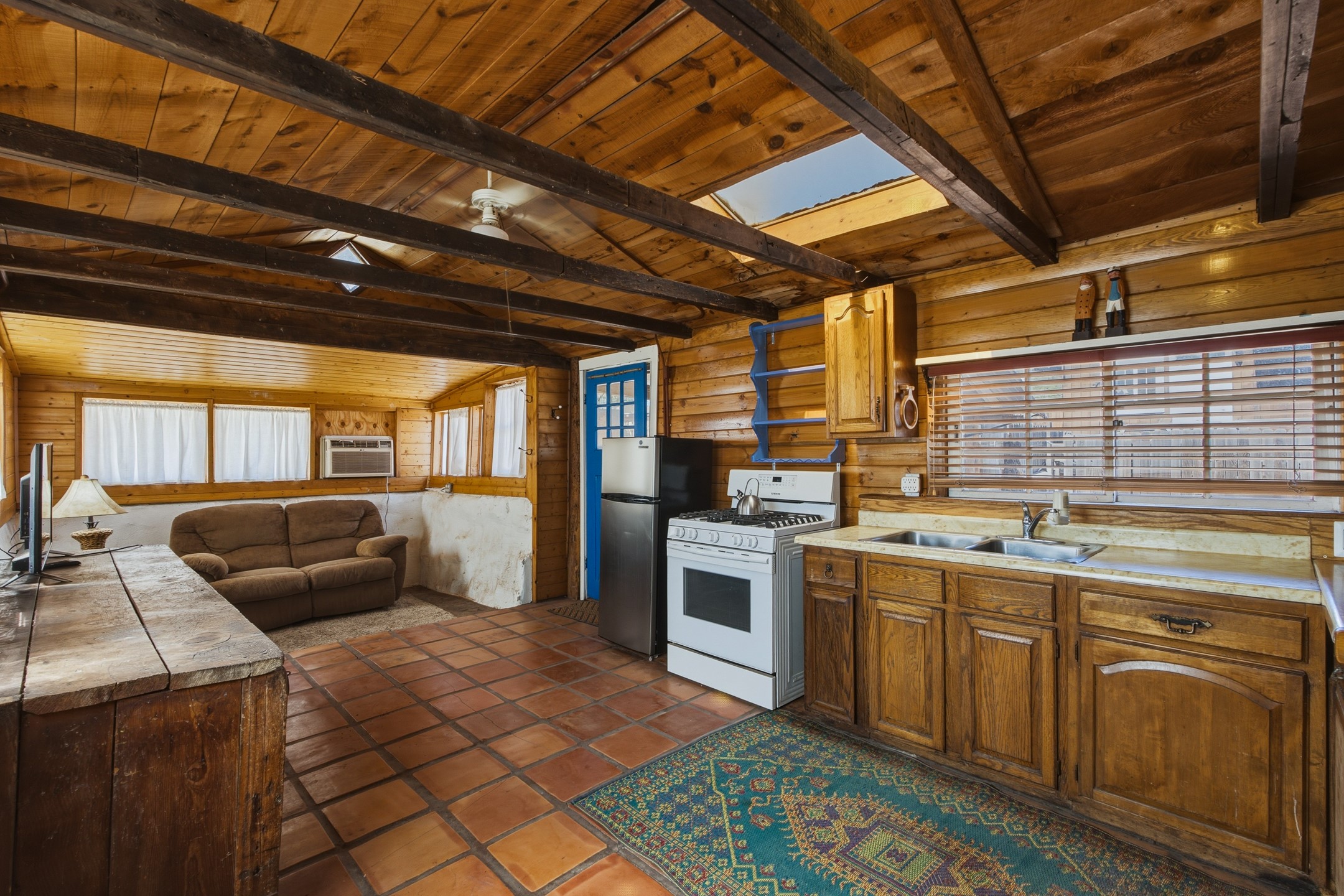 Wood-paneled kitchen and living area with exposed beams, skylight, terracotta floors, and brown sofa.