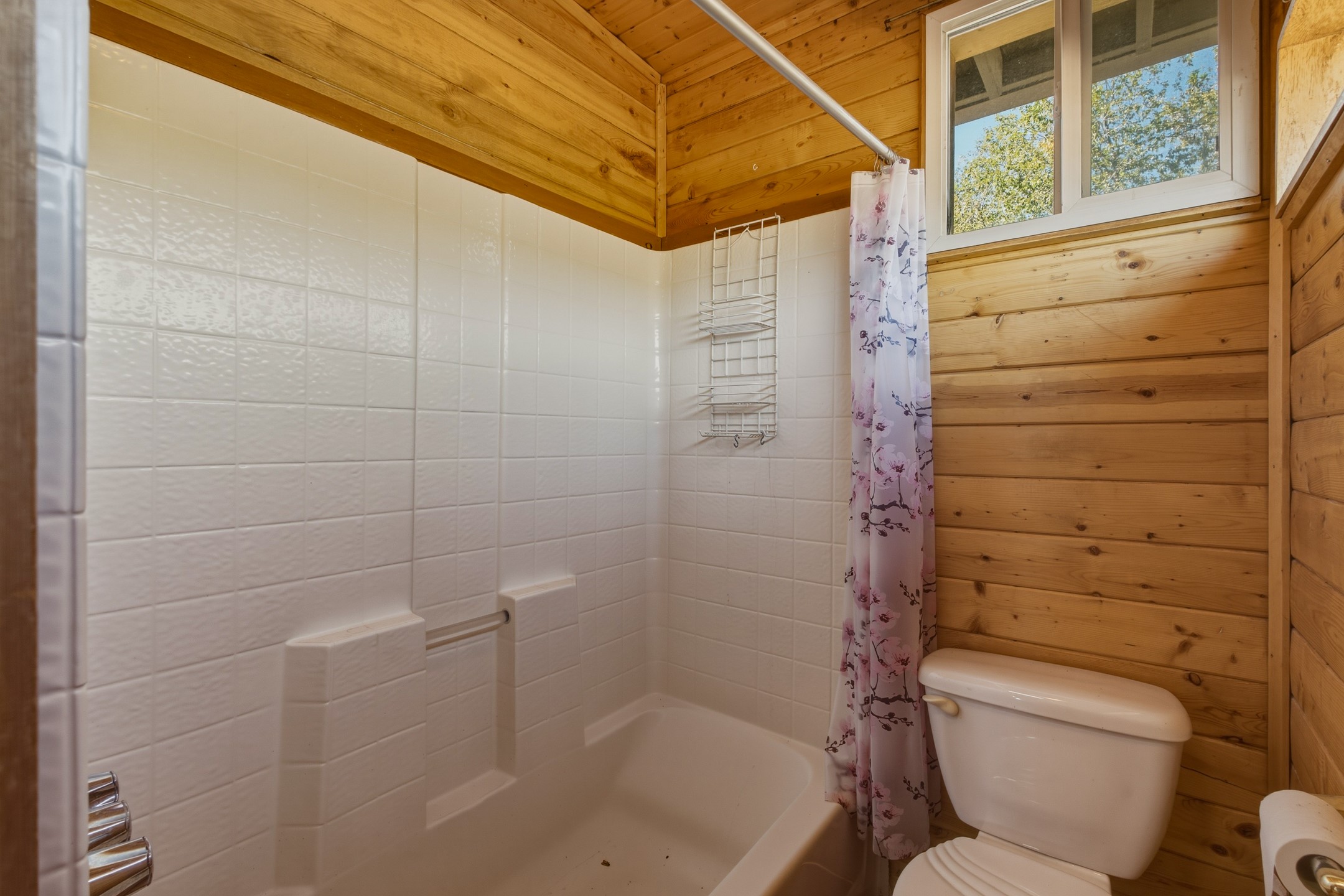 Bathroom with a white tiled tub, floral shower curtain, and wood-paneled walls.