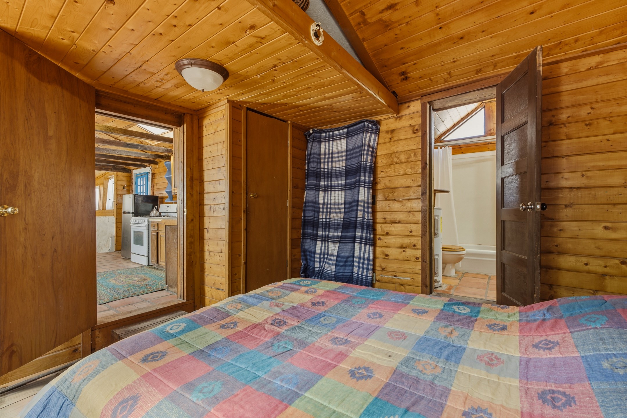 Rustic wood-paneled bedroom with a colorful patchwork bedspread, kitchen doorway, and bathroom doorway.